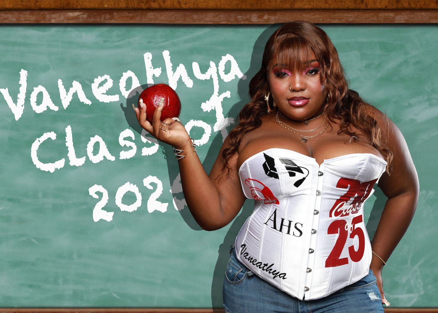 Graduate in customized outfit holds red apple against chalkboard background.