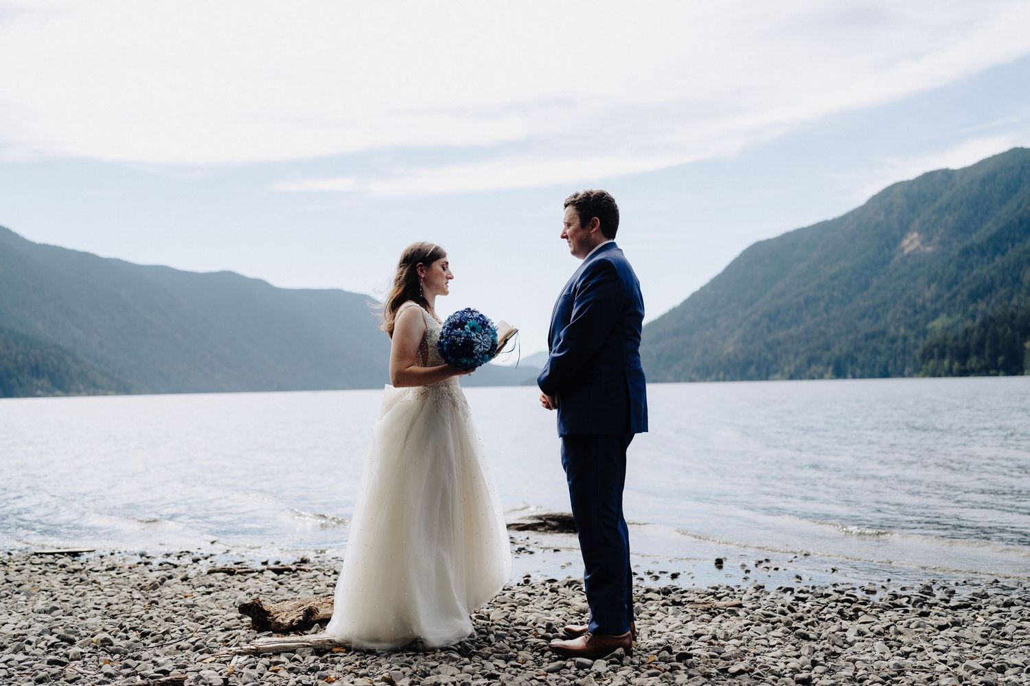 A wedding couple stands on a rocky beach with mountains and lake in the background on their special day.