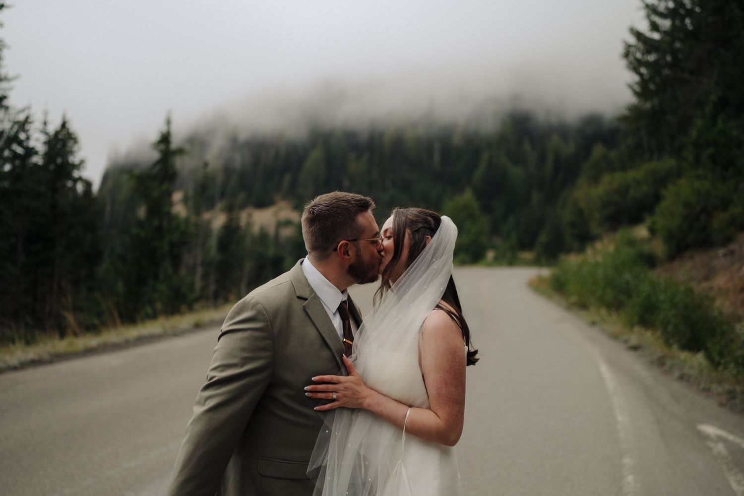 A couple shares a romantic kiss on a misty mountain road surrounded by evergreen forest.