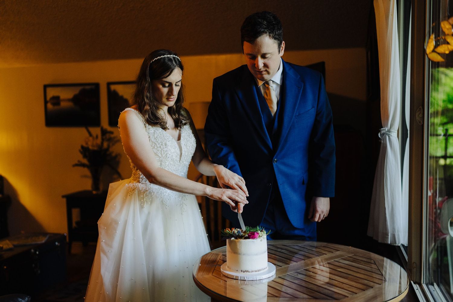 A couple cuts their wedding cake together in a warmly lit indoor setting.