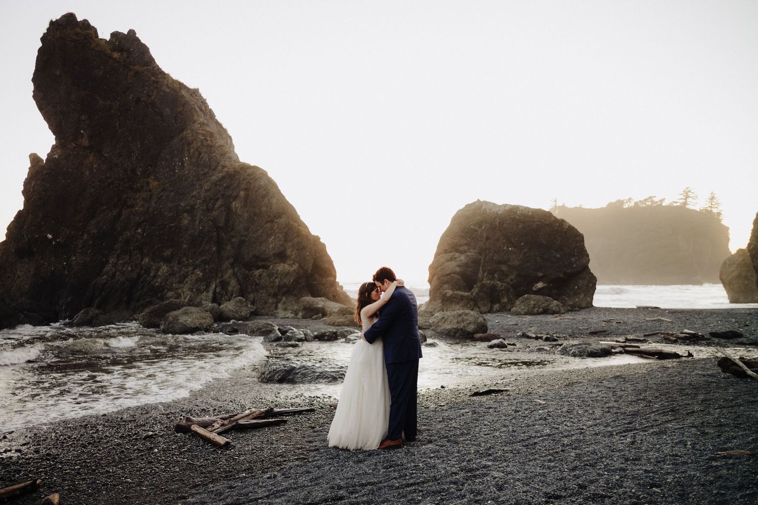A couple embraces on a rocky beach with dramatic sea stacks and misty coastline in the background.