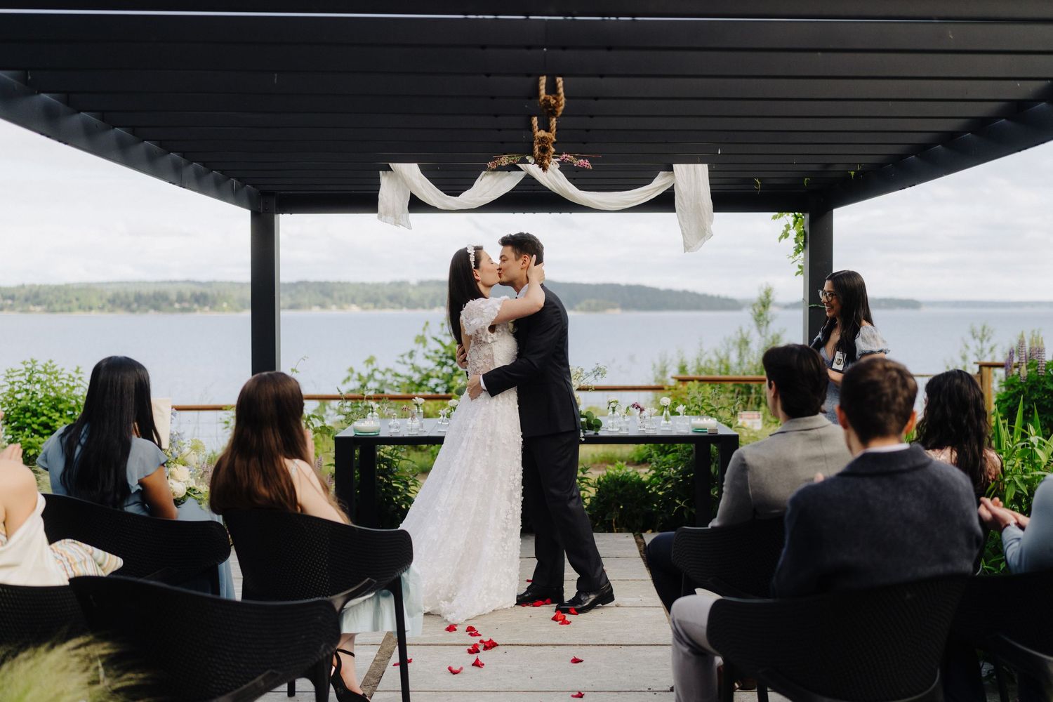 A couple shares a romantic moment during their lakeside wedding ceremony as guests look on from covered seating area.