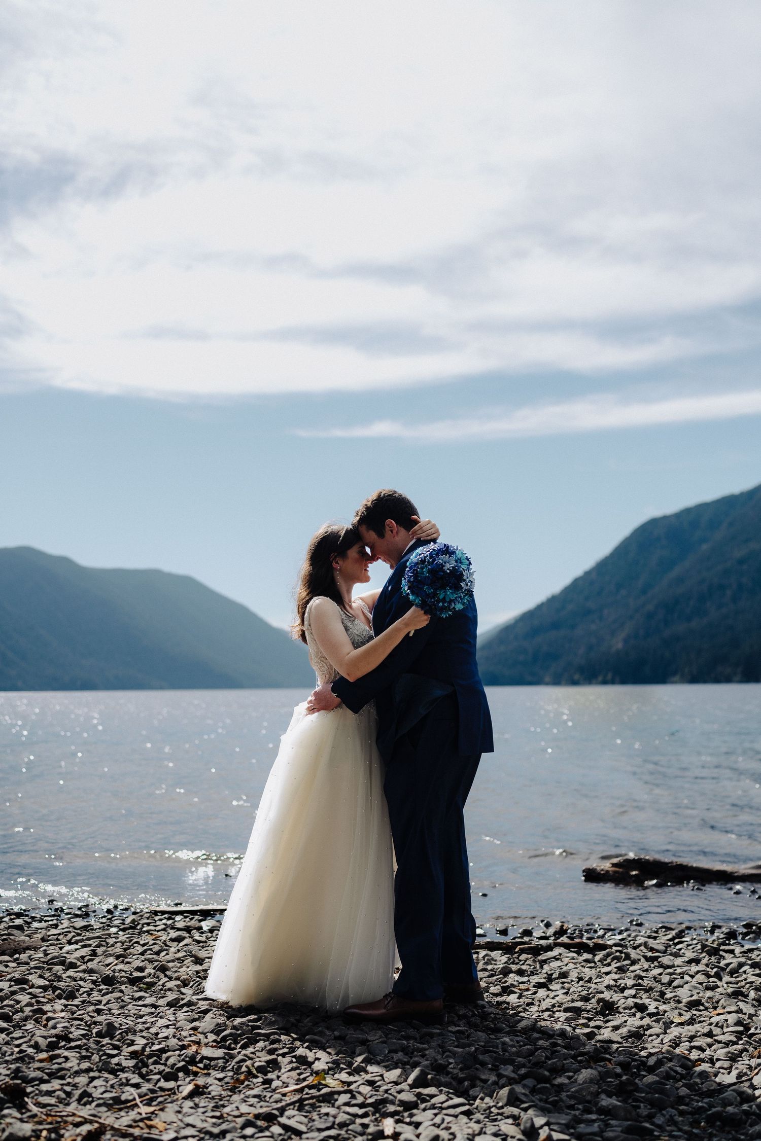 Intimate wedding moment captured on pebbly shore with sunlit mountain lake backdrop.