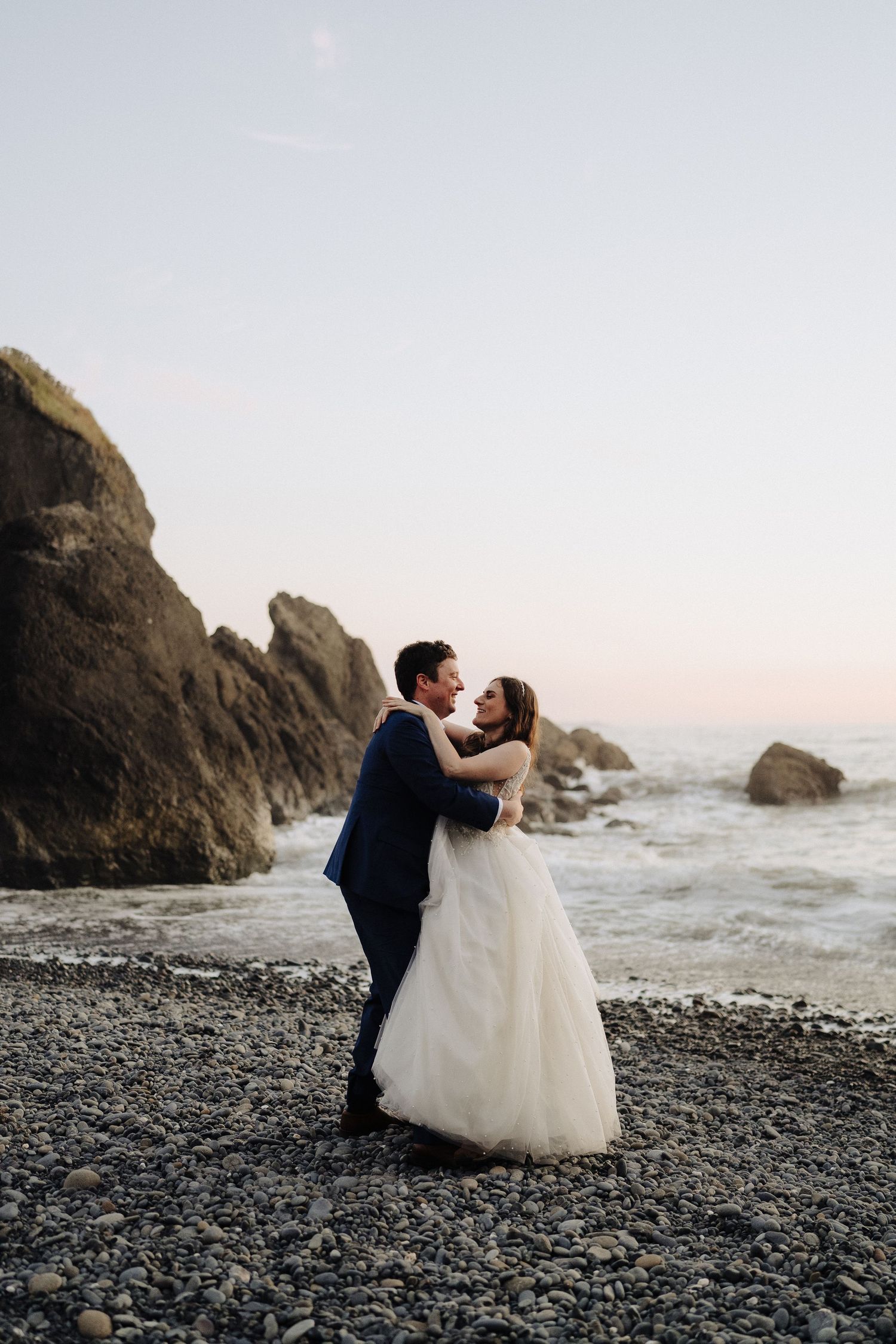 Newlyweds embrace on a rocky beach with dramatic coastal cliffs in the background at sunset.
