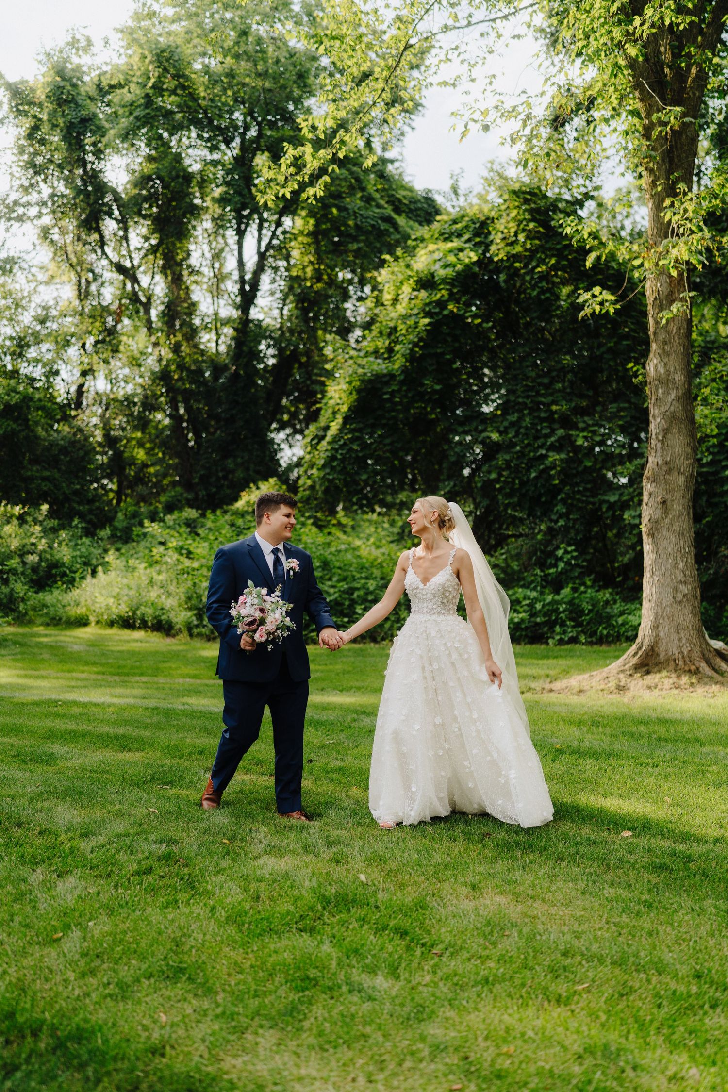 Newlyweds walking together on green lawn surrounded by tall trees.