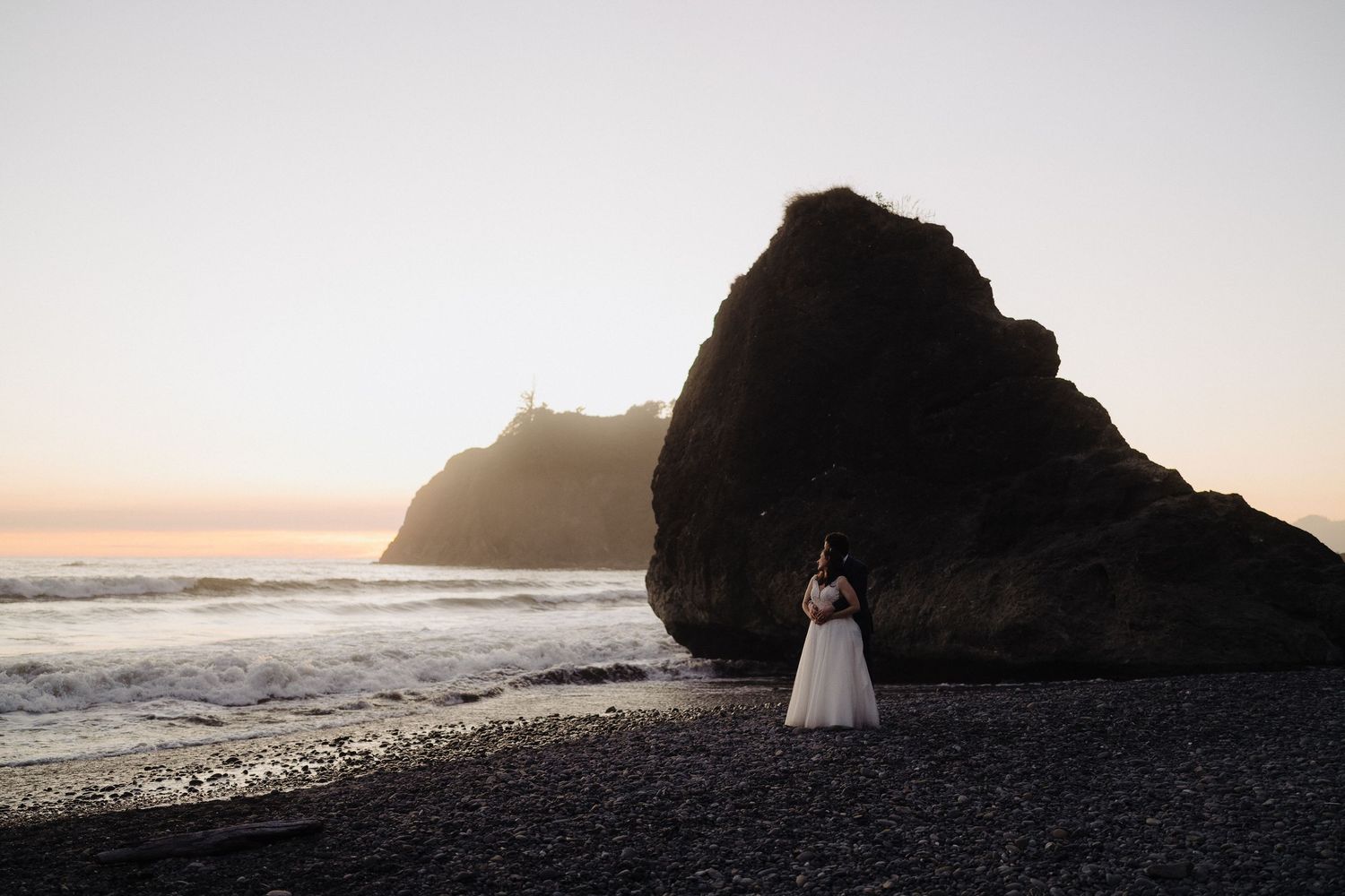 A white dress silhouette against dramatic coastal rocks during sunset at the beach.