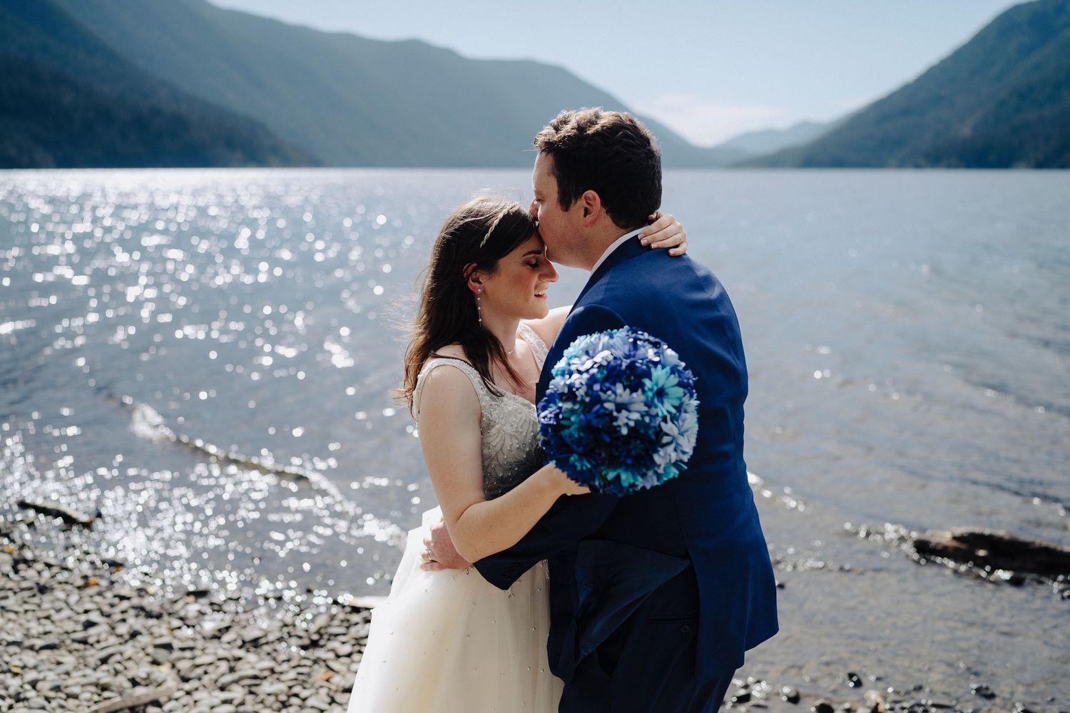 Tender moment between newlyweds with sparkling lake and mountain landscape.