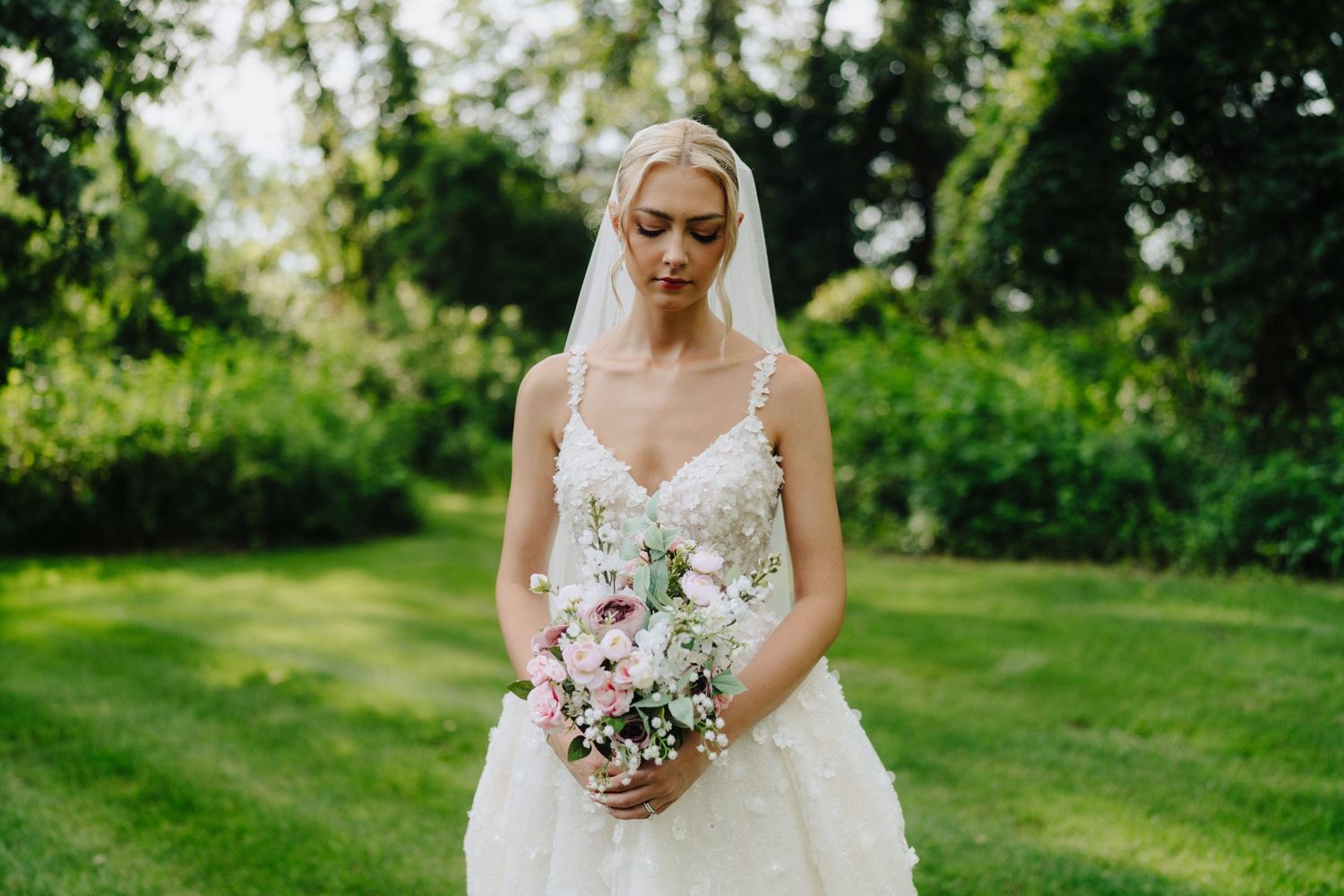 Bride in lace wedding dress holding pink and white bouquet in garden setting.