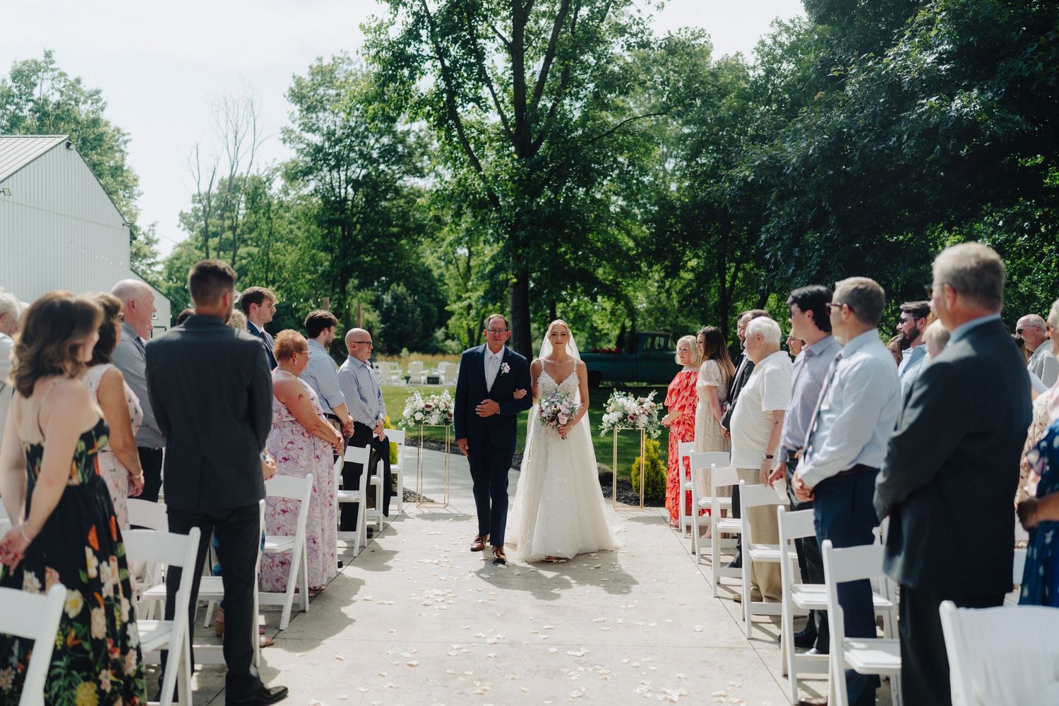 Outdoor wedding ceremony with guests seated in white chairs along a tree-lined path.