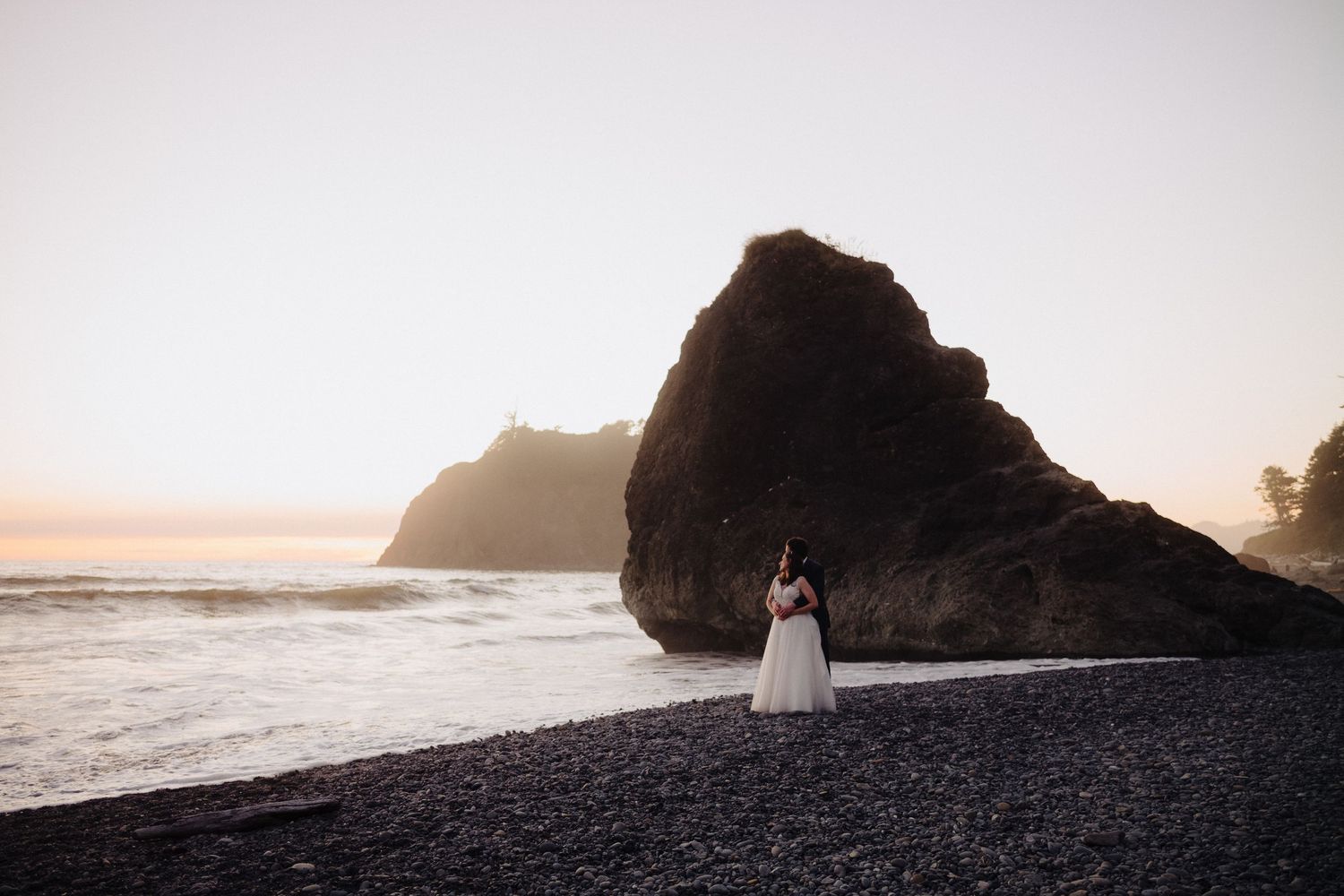 Two silhouettes embrace on a rocky black sand beach at sunset with dramatic coastal cliffs in the background.