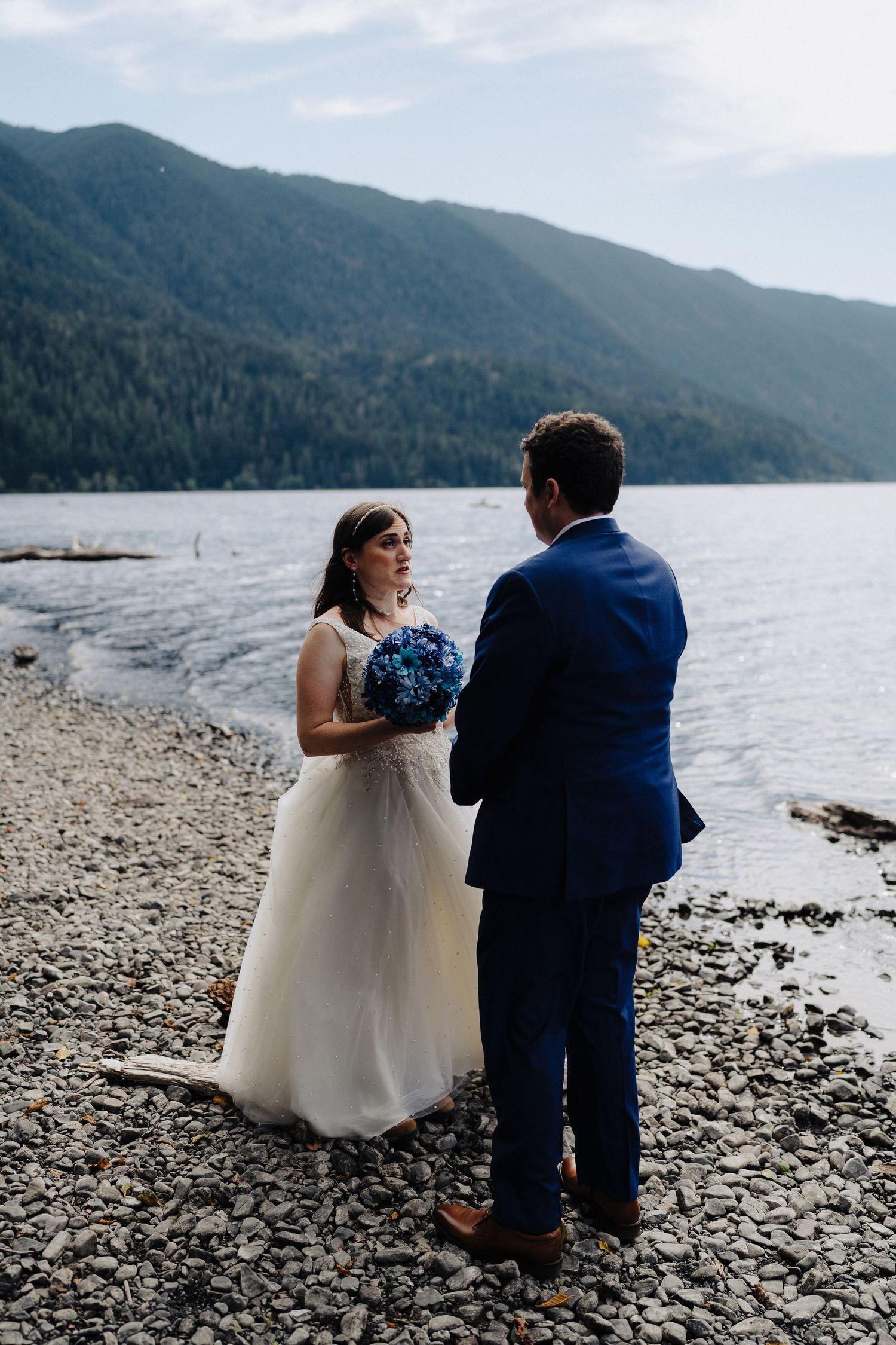 A wedding couple stands on a rocky beach shore next to a mountain lake holding blue flowers.