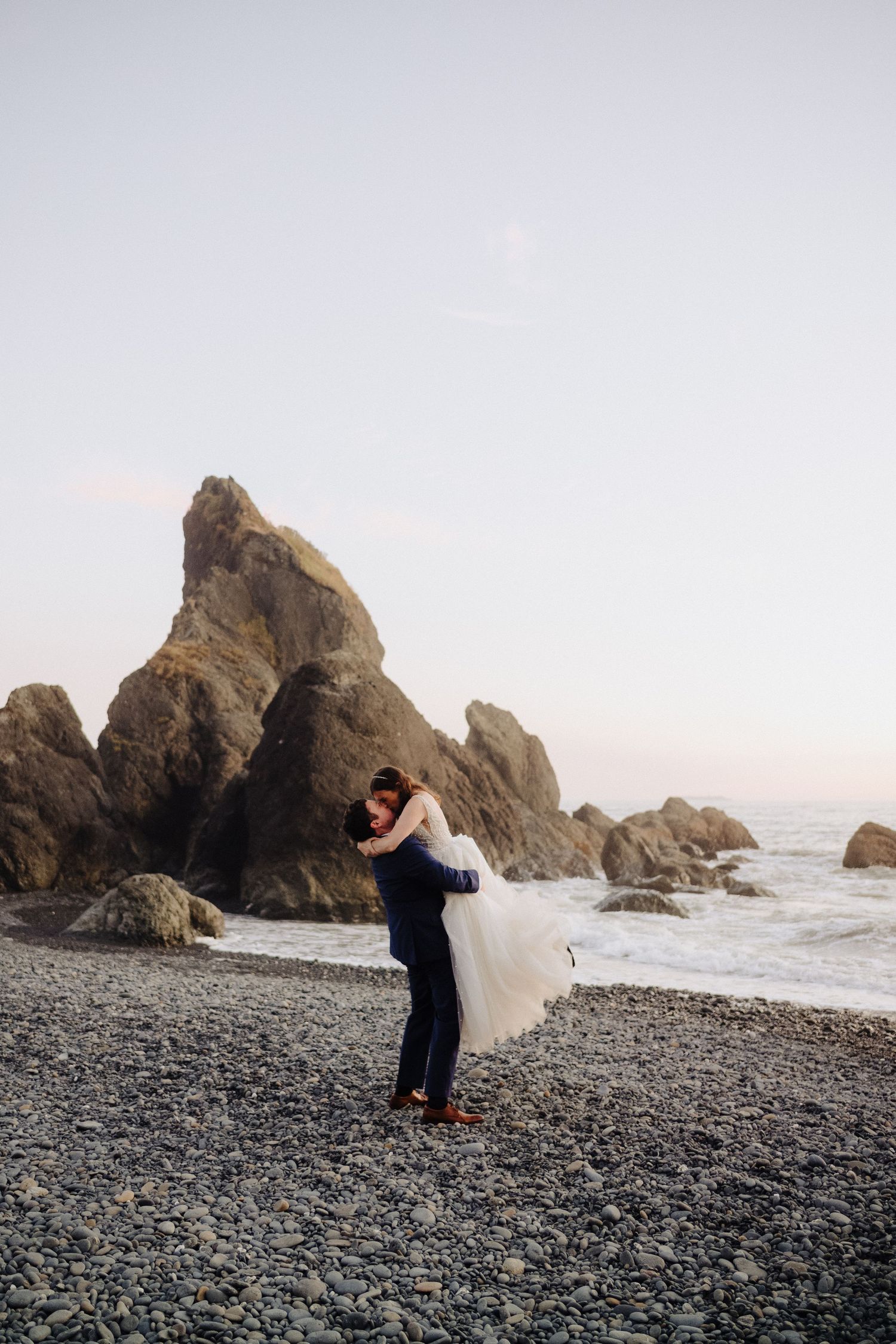 Couple in wedding attire embracing on a rocky beach with dramatic coastal rock formations behind them.