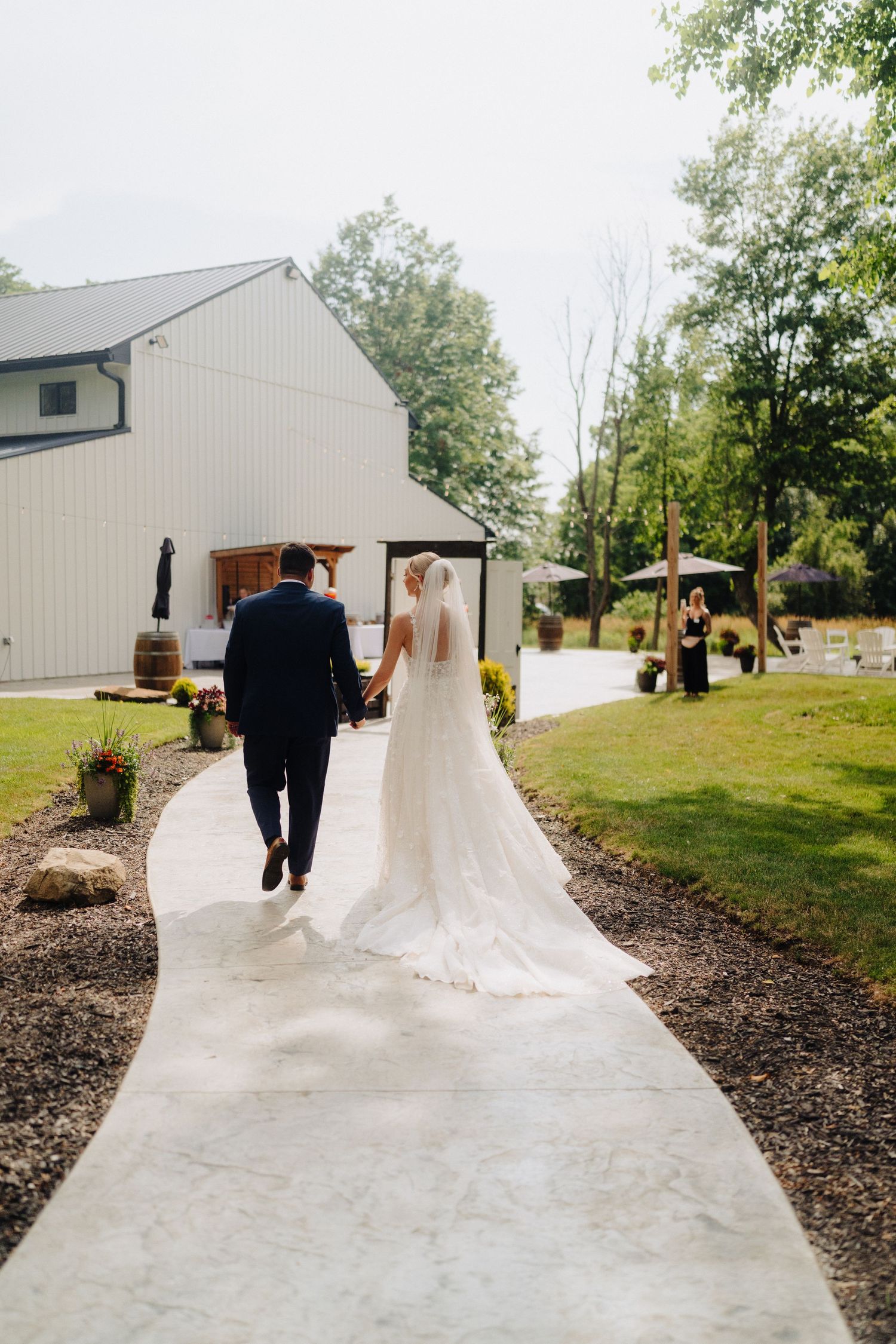 A couple walks along a concrete path outside a white barn-style building on their wedding day.