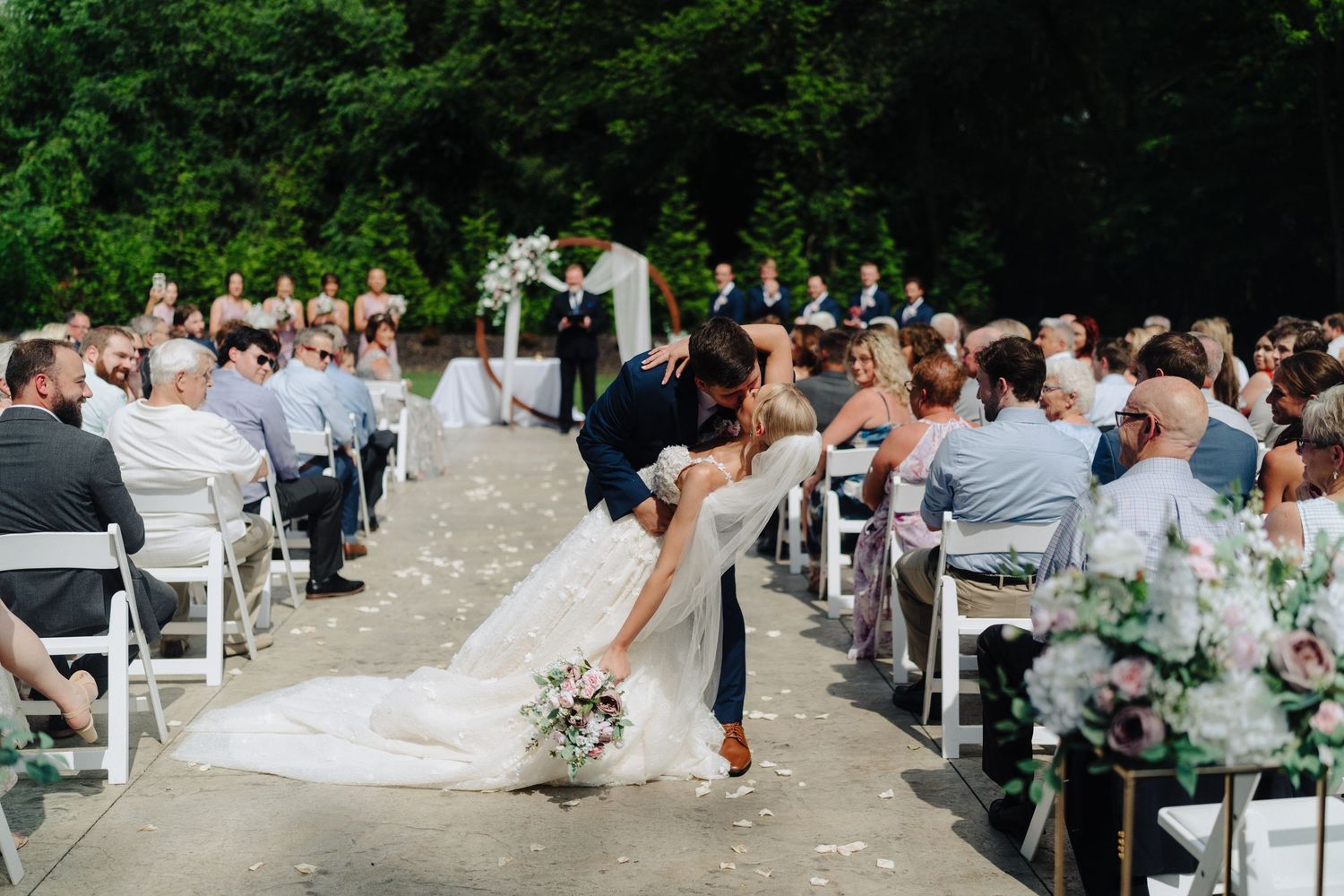 Panoramic view of wedding guests gathered for an outdoor ceremony with white chairs and greenery.