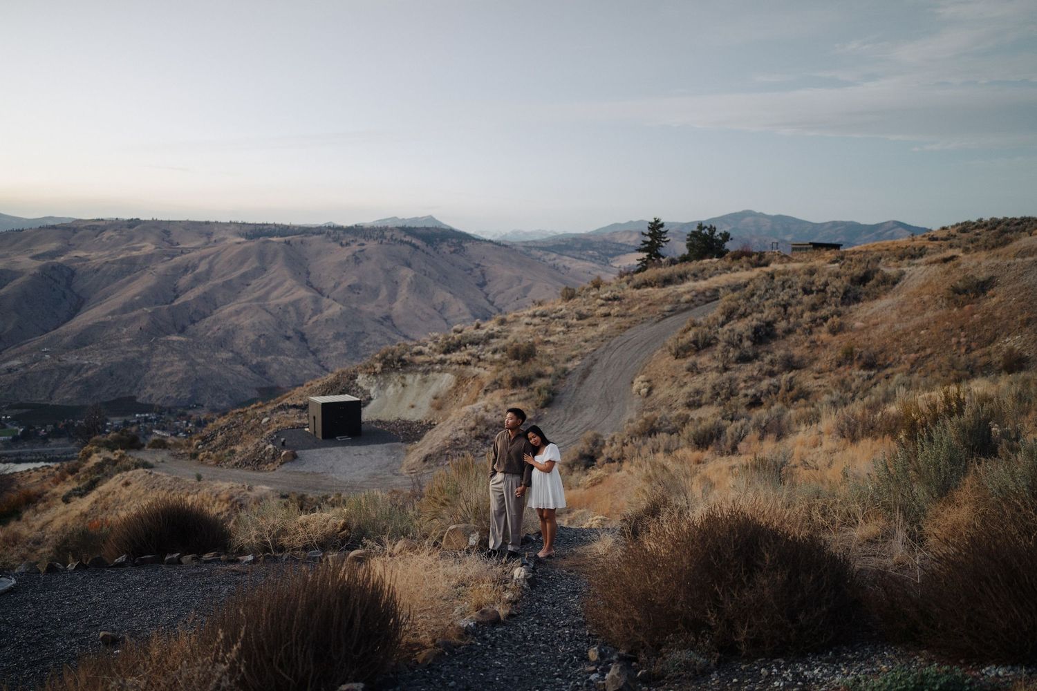 A scenic mountain trail leads through rugged terrain with distant peaks and scrubland vegetation in the background.