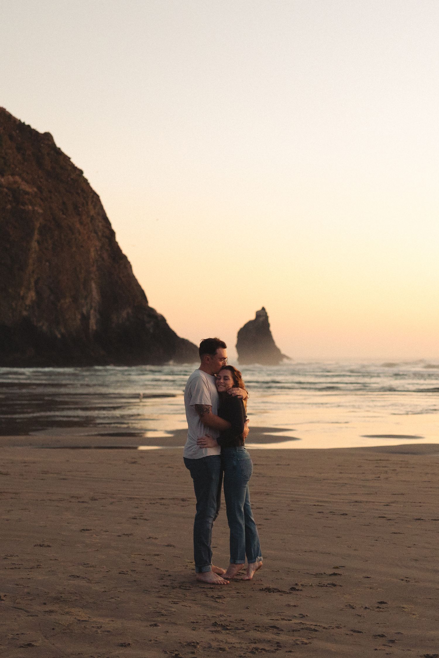Couple embracing on beach at sunset with dramatic coastal rock formations in background.