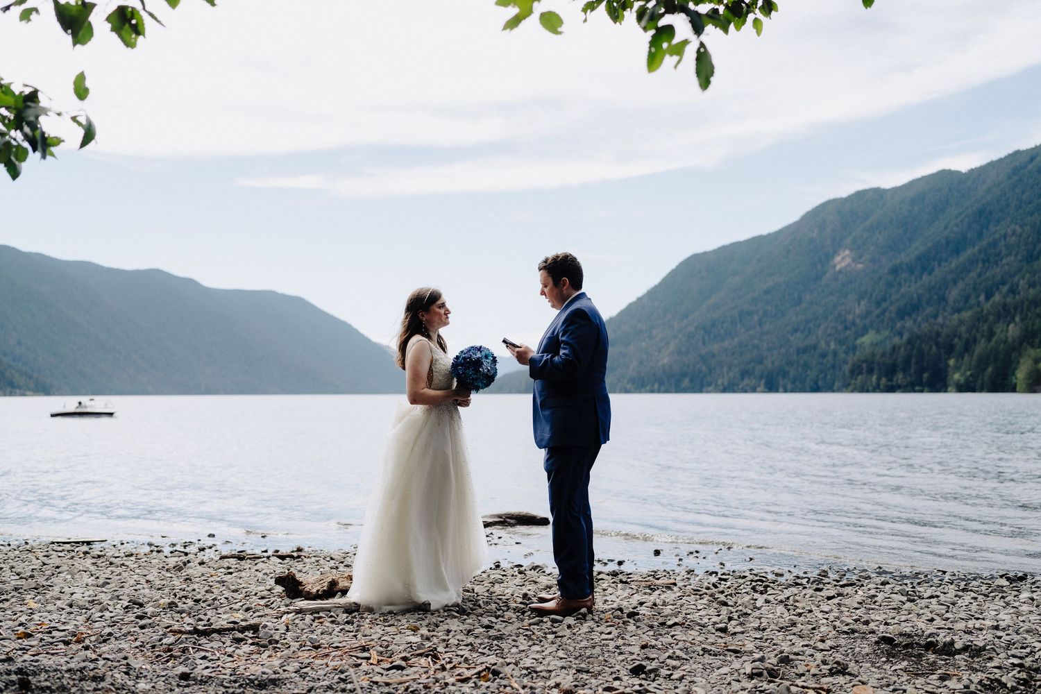 A couple exchanges wedding vows on a rocky beach with mountain lake views and forested hills in the background.