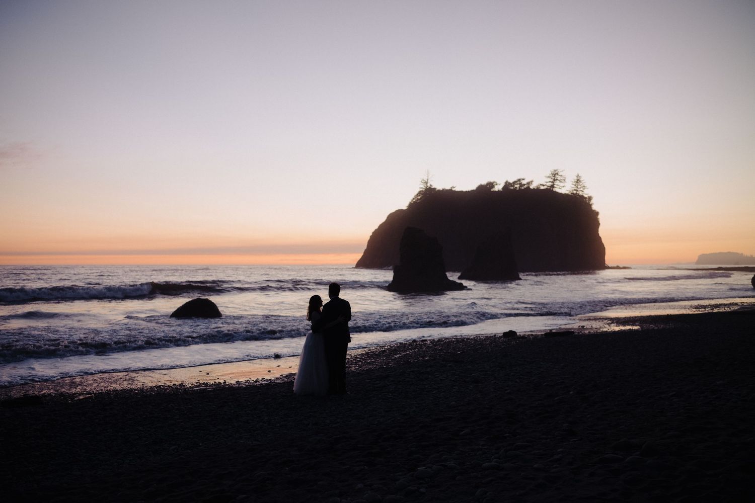 Silhouette of a couple standing on a beach with a large rock formation at sunset.