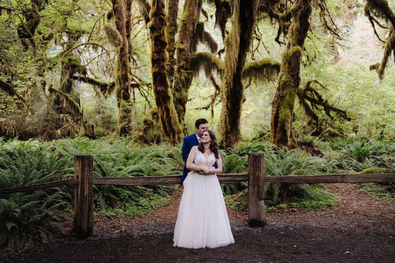 A wedding couple embraces on a wooden boardwalk in a lush mossy forest.