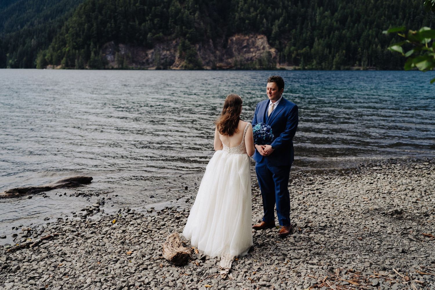 A romantic wedding photoshoot with a couple standing by a scenic mountain lake on a rocky shore.