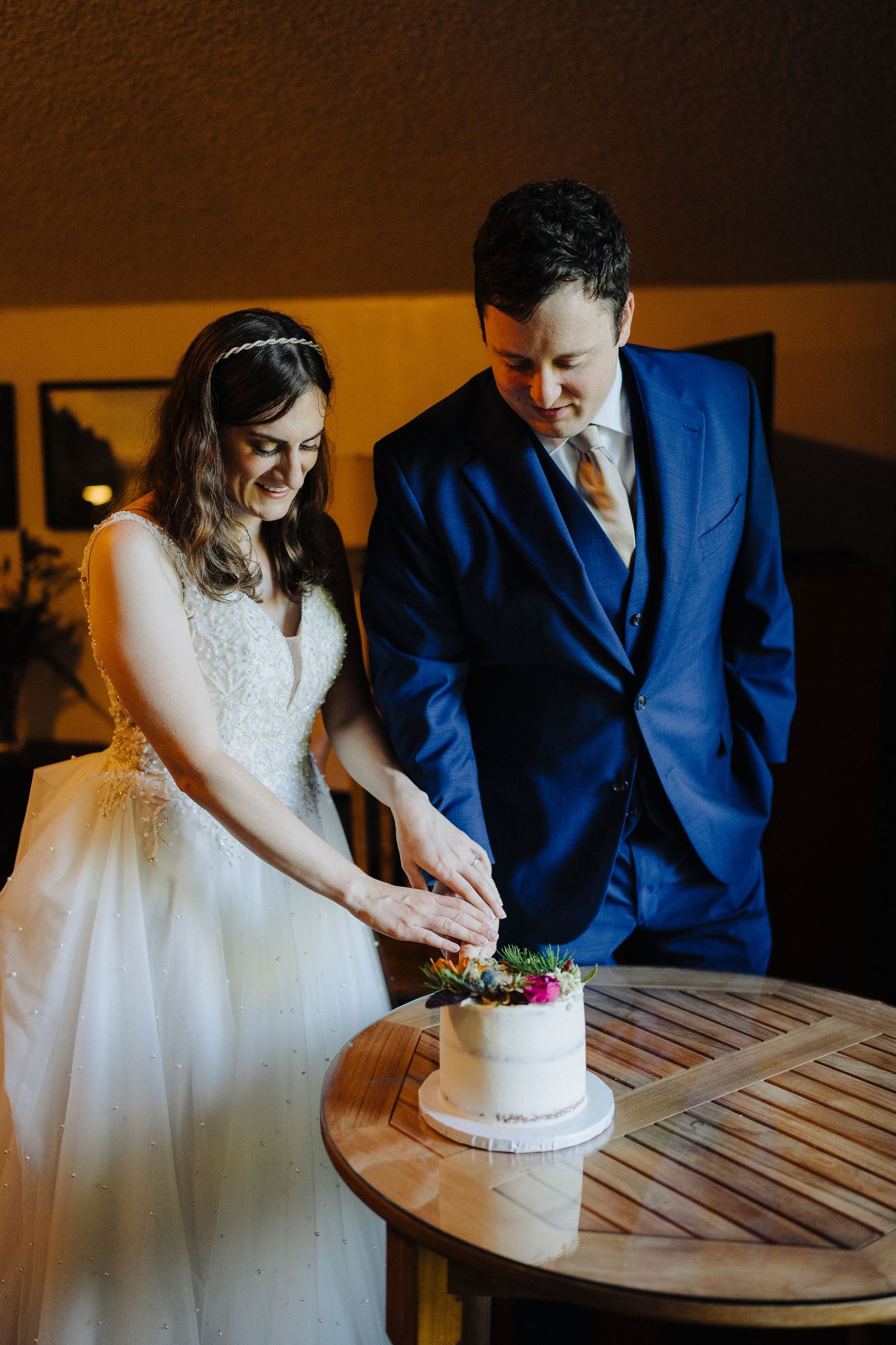 A couple cuts their simple white wedding cake together at a wooden table indoors.