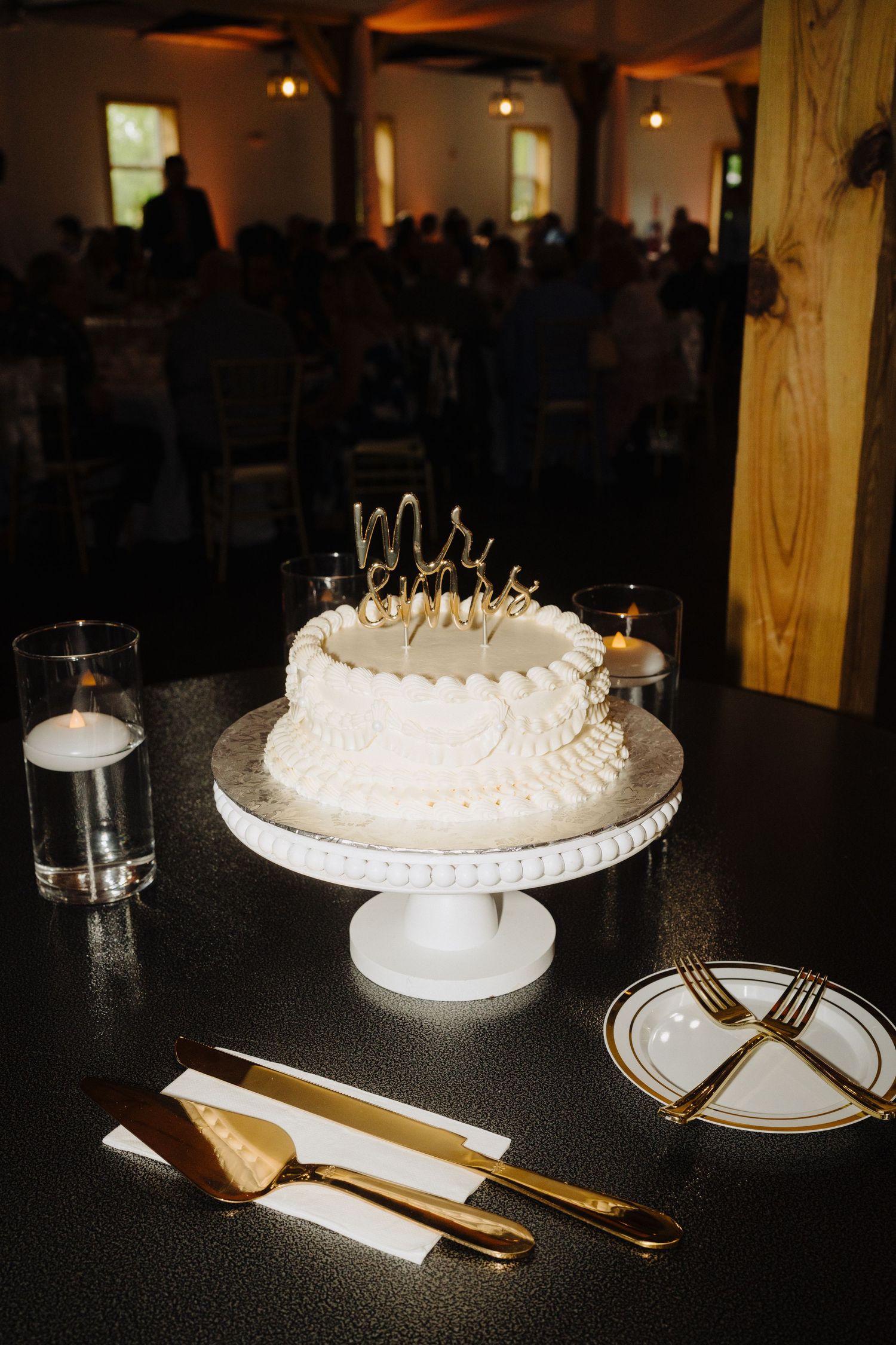 A white wedding cake with delicate piping sits on a black table with gold utensils and place settings.