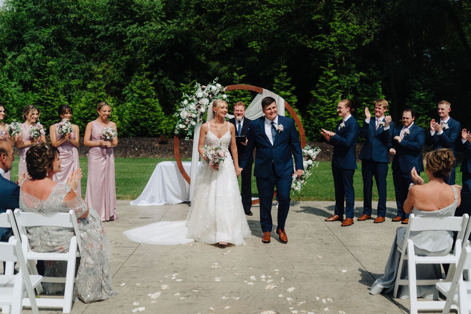 Wedding ceremony with bridal party and guests seated on white chairs under a floral arch outdoors.