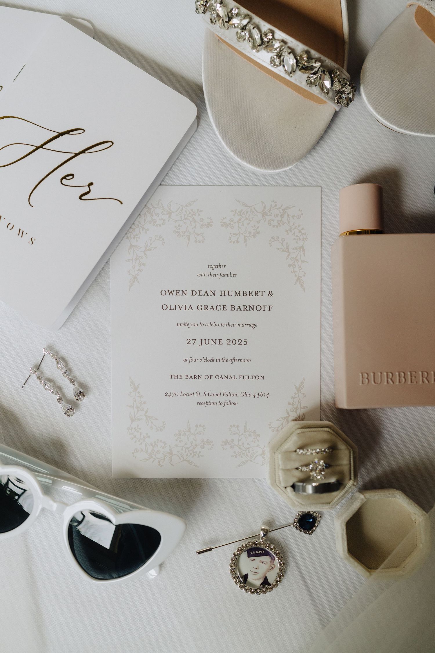 Wedding invitation and bridal accessories including sunglasses, jewelry, and a Burberry perfume on white surface.