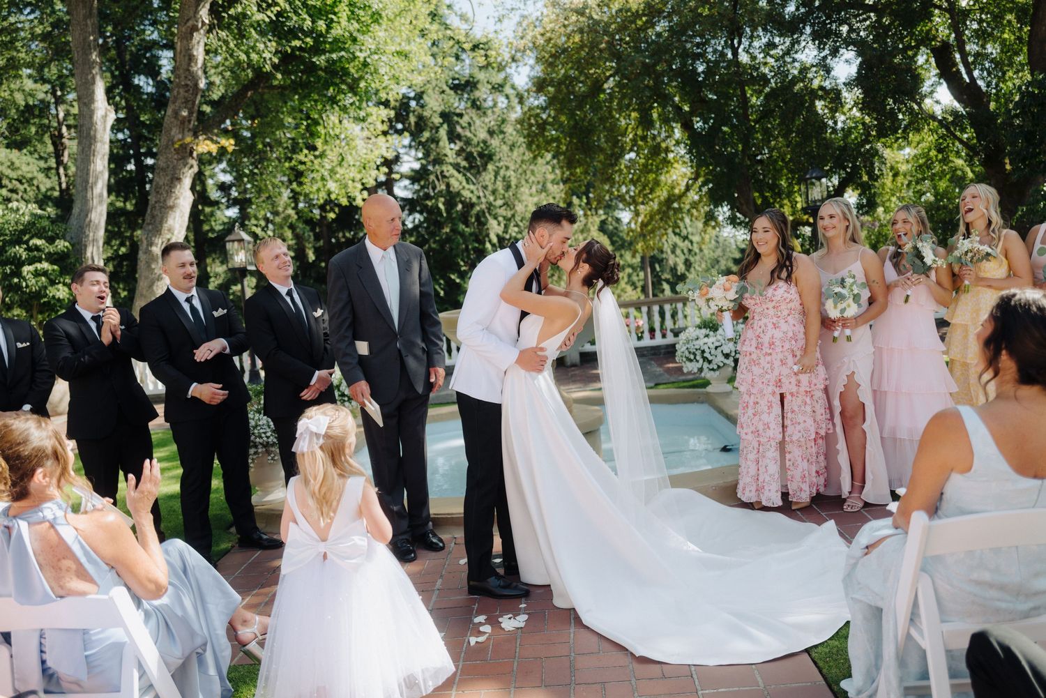Elegant outdoor garden wedding ceremony with couple kissing while guests in pink and blue dresses look on.