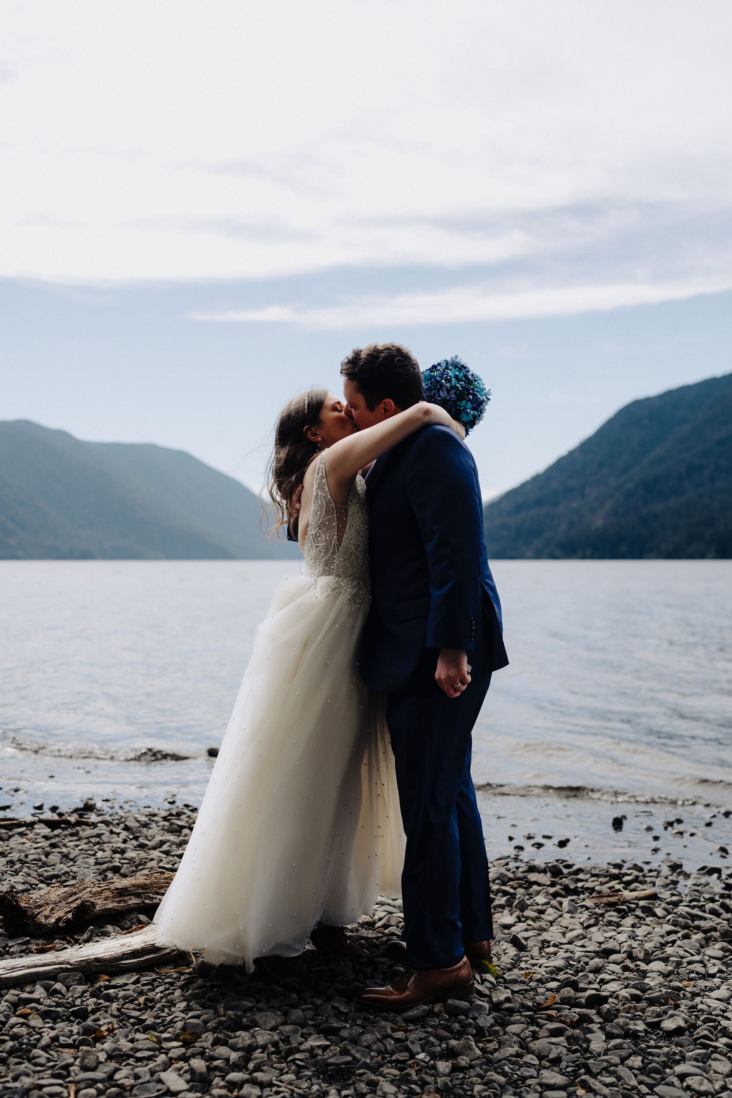 Romantic wedding couple embraces on lakeside beach with mountain vista behind them.