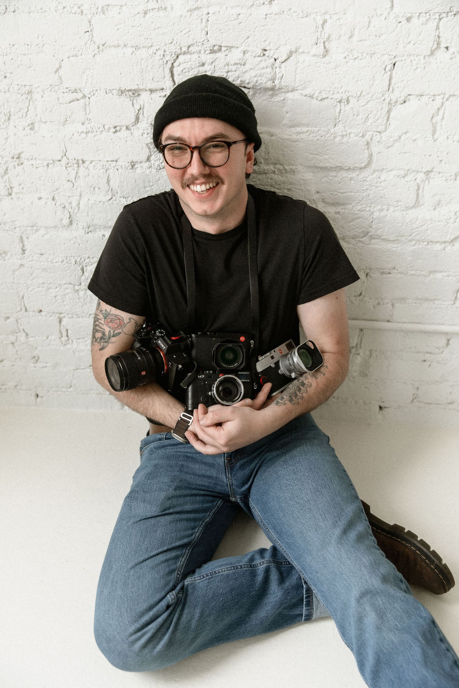 Photographer in black beanie and t-shirt sits against white brick wall holding vintage film cameras.