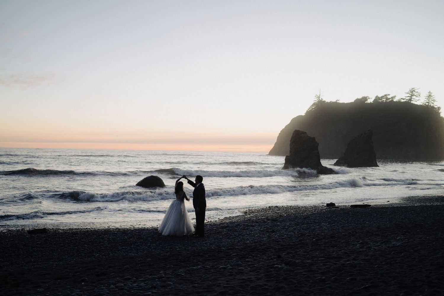 Silhouettes stand against a sunset beach scene with rocky cliffs in the background.