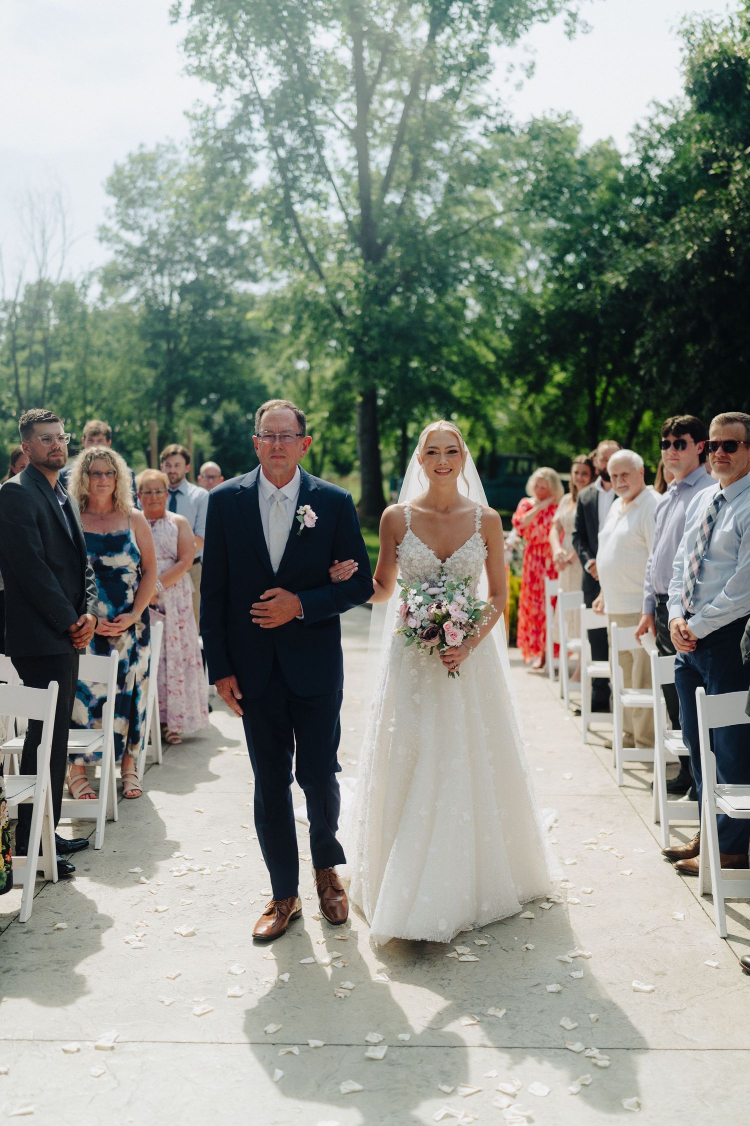 Wedding ceremony guests line white chairs on an outdoor path surrounded by lush greenery on a sunny day.