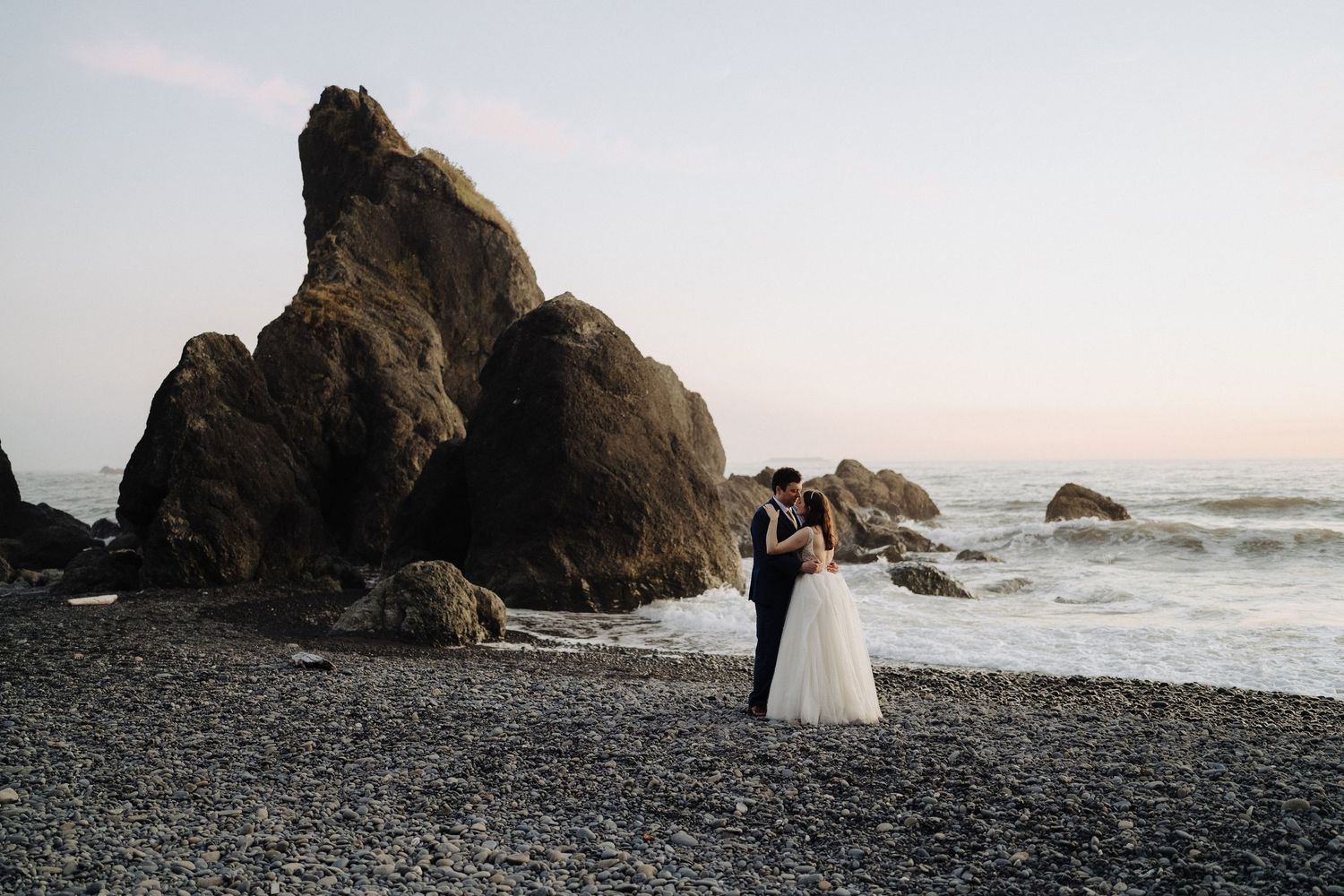 Wedding couple embraces near dramatic coastal rock formations at sunset.