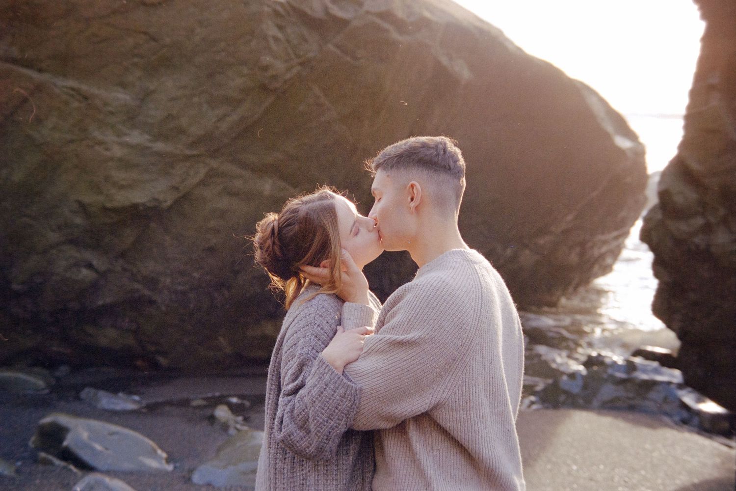A couple shares a romantic moment on a beach at sunset with dramatic rock formations in the background.