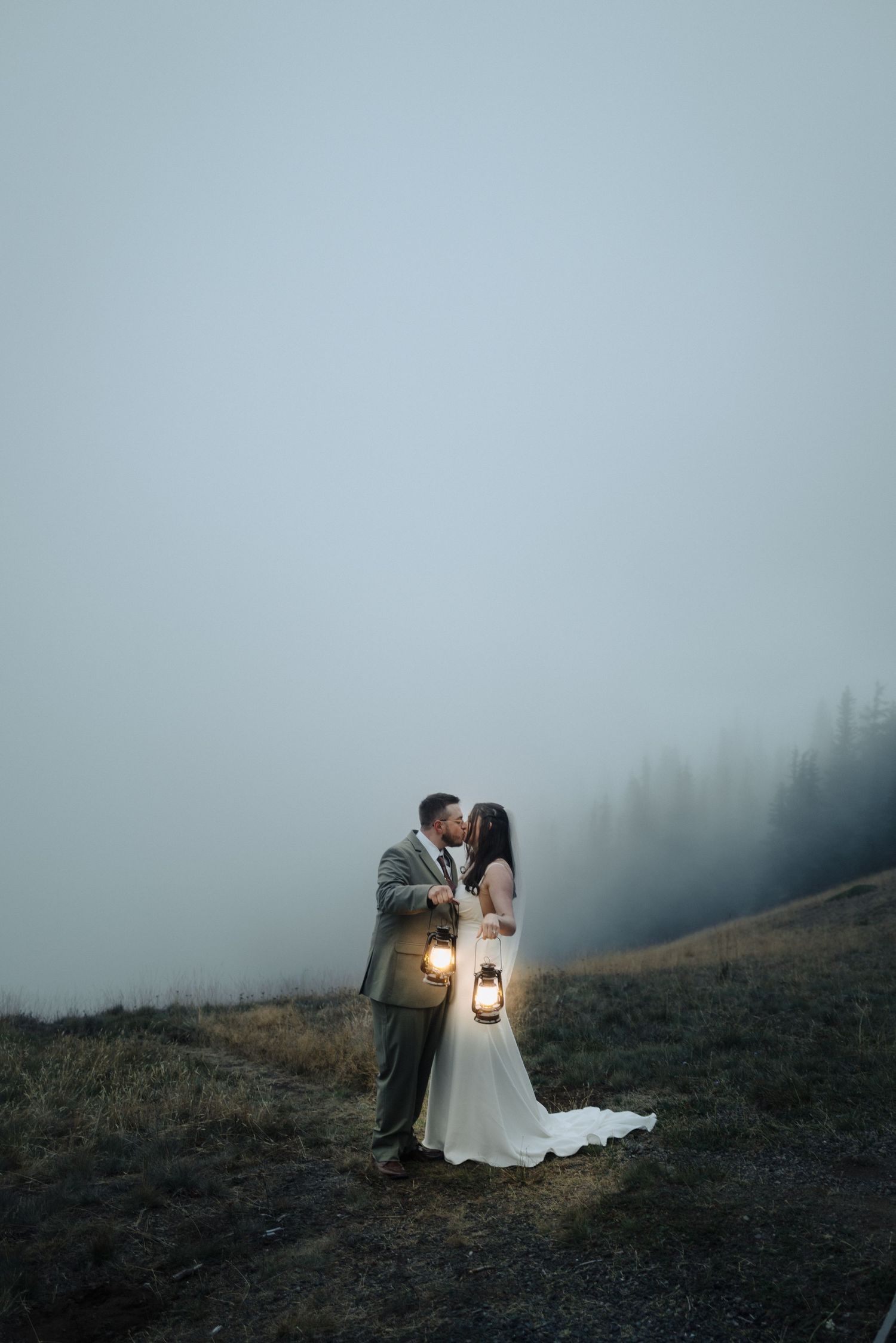 A romantic couple in wedding attire holds lanterns on a misty mountainside at dusk creating a magical atmosphere.