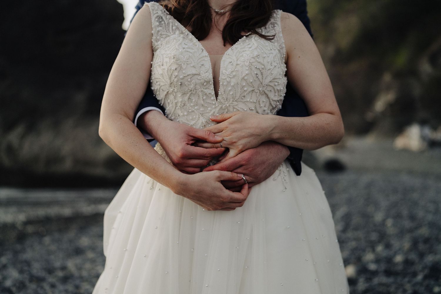 A detail shot of hands resting on a white wedding dress against a rocky beach backdrop.