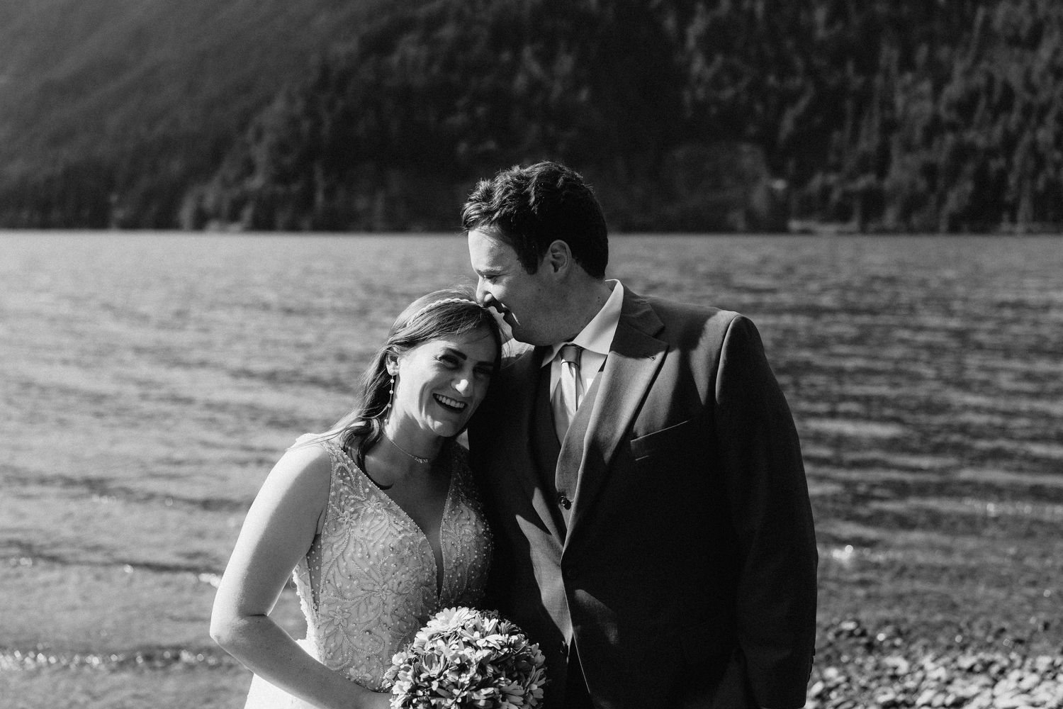 A black and white portrait of a couple sharing an intimate moment on a beach with mountains behind them.