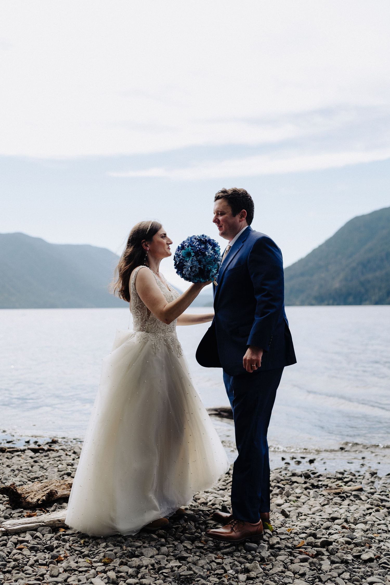 Wedding couple poses playfully on rocky beach with mountain views.