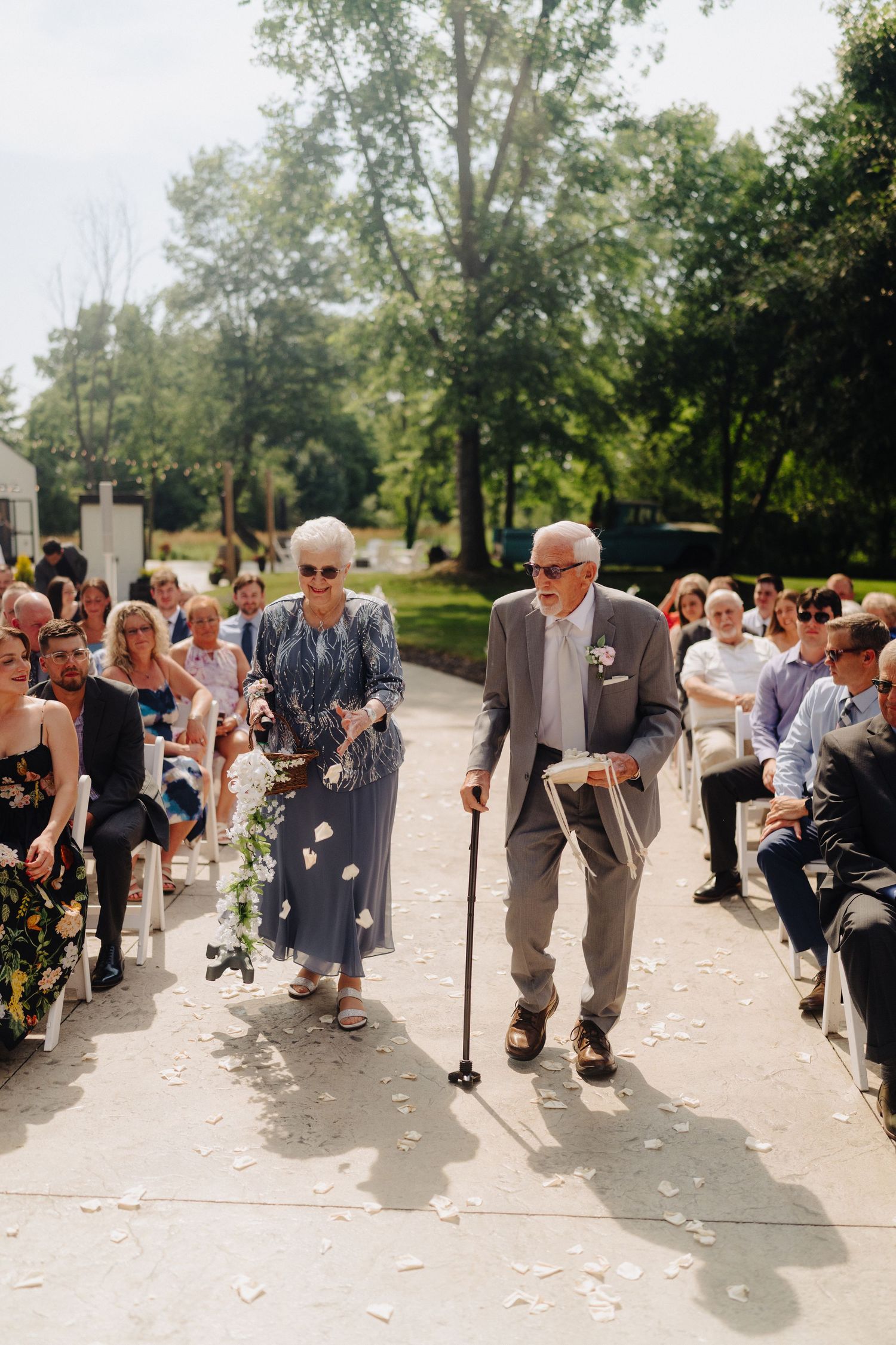 Wedding ceremony guests seated outdoors on a sunny day with tree-lined pathway.