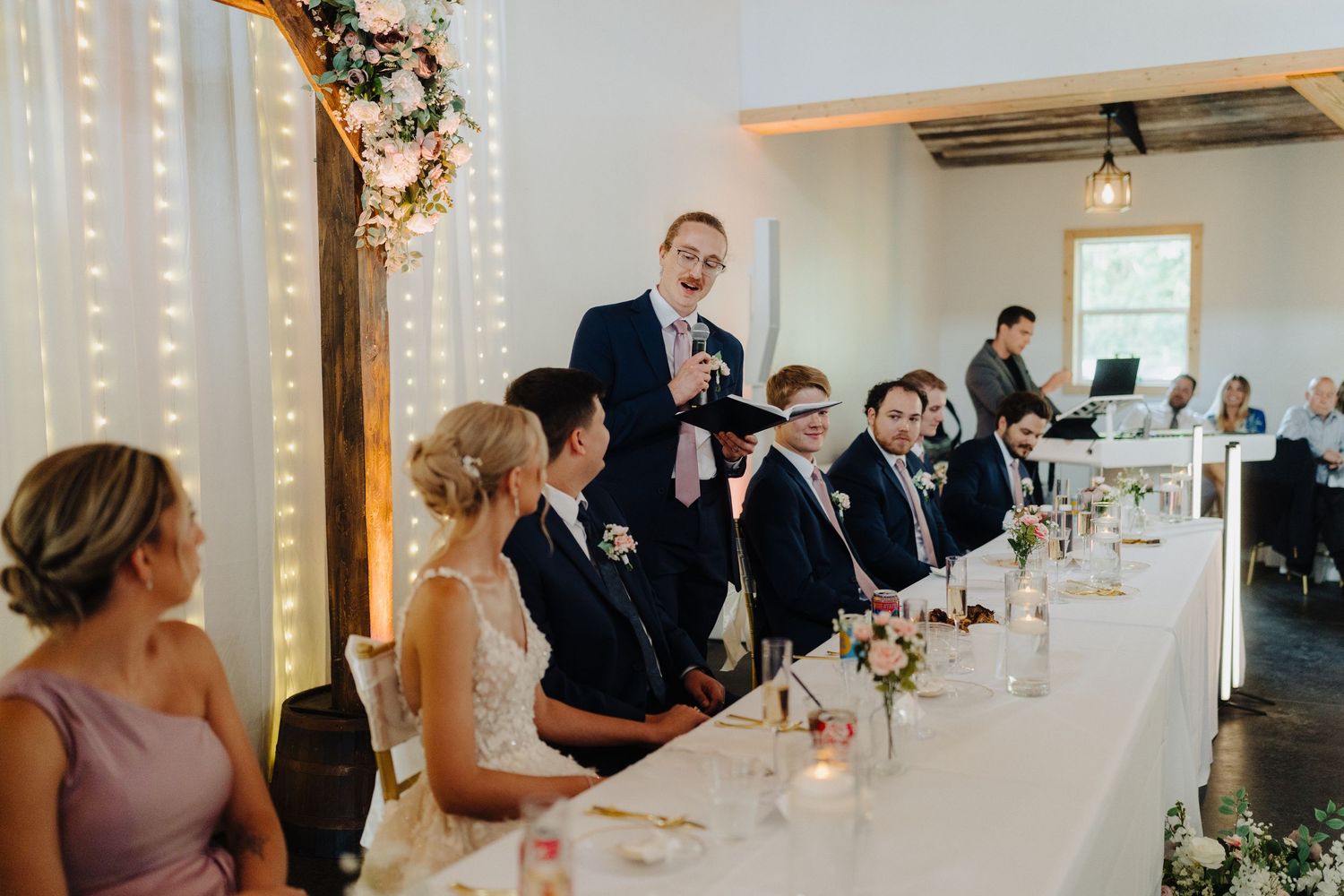 Wedding reception guests seated at elegant long tables with candlelight and floral centerpieces in a bright venue.