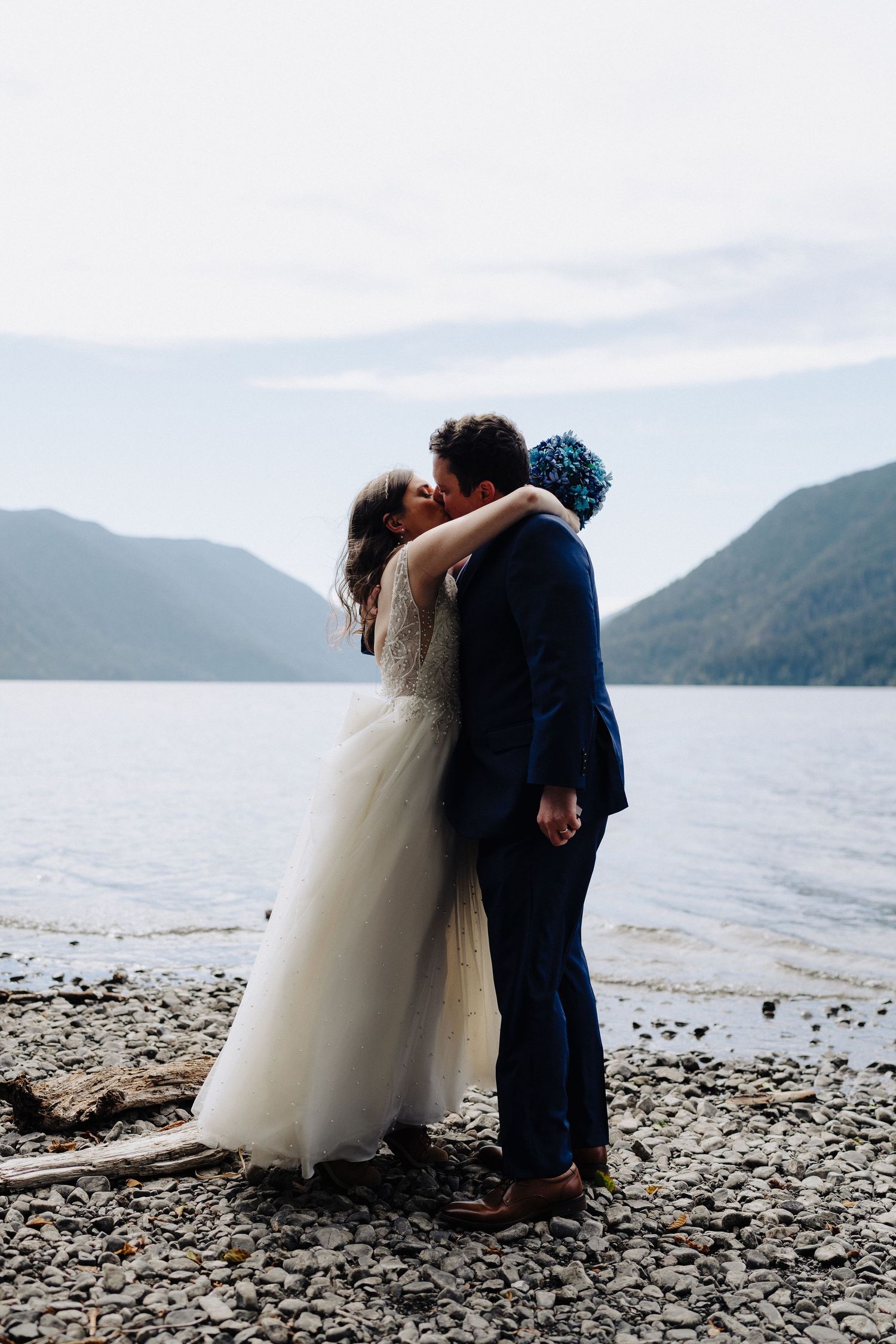 Couple in wedding attire sharing intimate moments by a serene lakeside with mountains in the background.