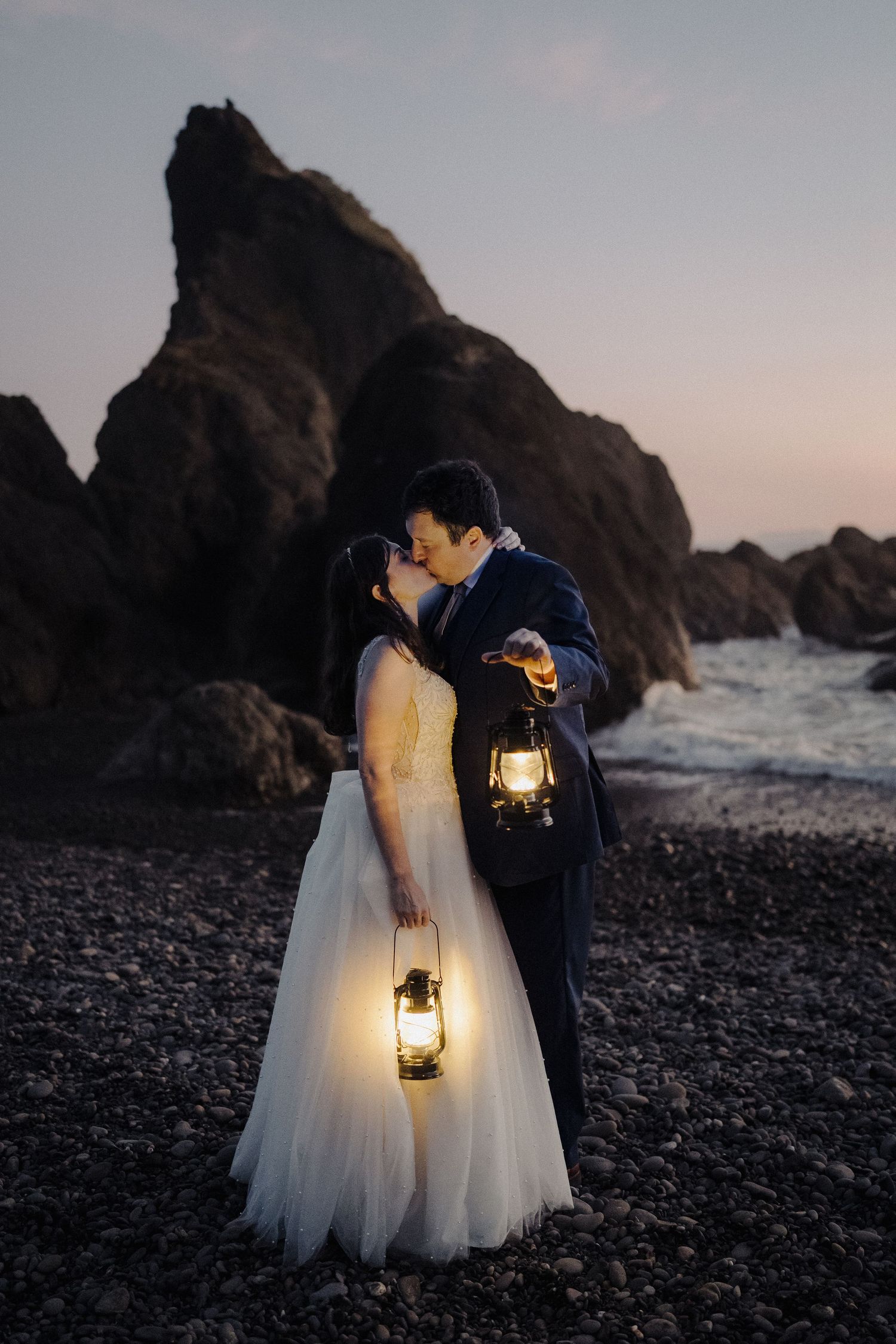 Couple with lanterns shares romantic moment on rocky beach at dusk.