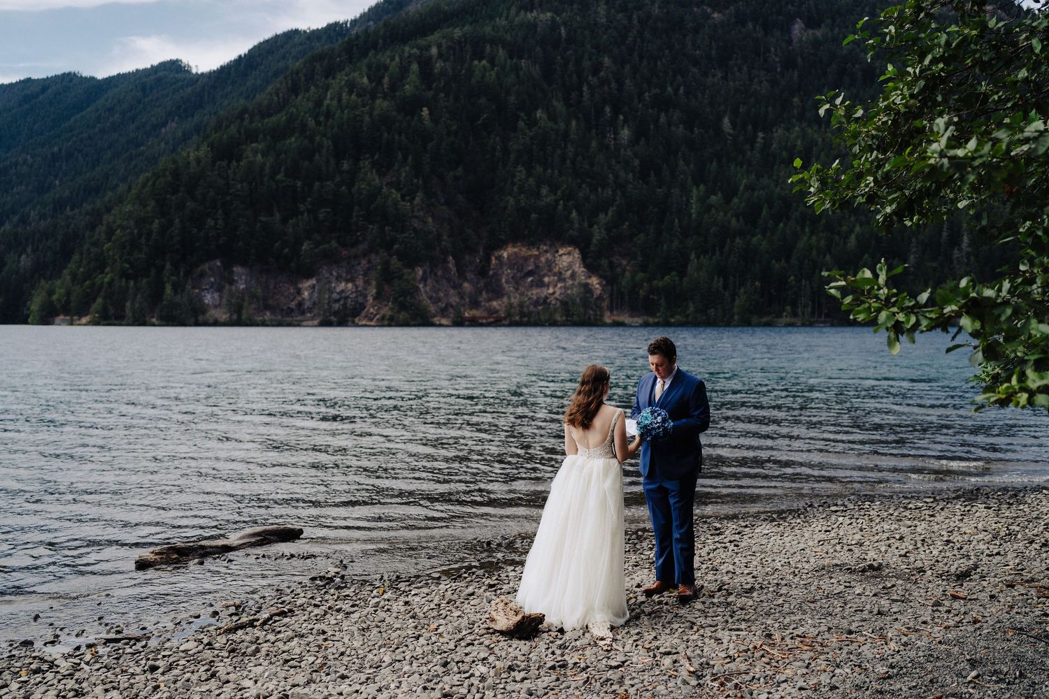 Wedding couple shares a moment on a pebble beach surrounded by mountains and lake views.