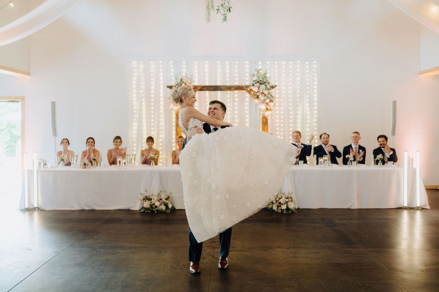 Panoramic view of a modern white wedding reception hall with head table and string light decorations.