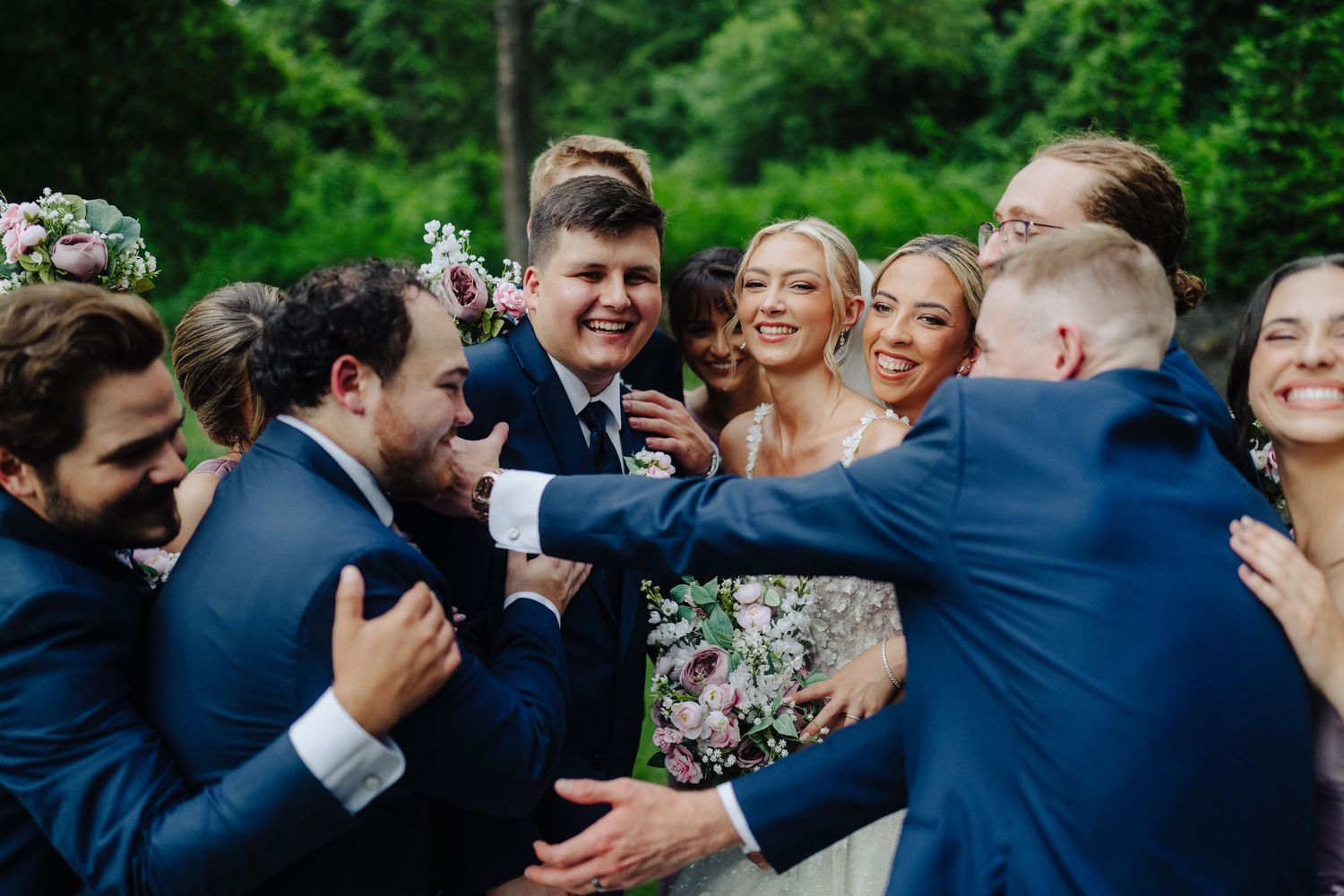Wedding party celebrates with joyful hugs and laughter in a garden setting.