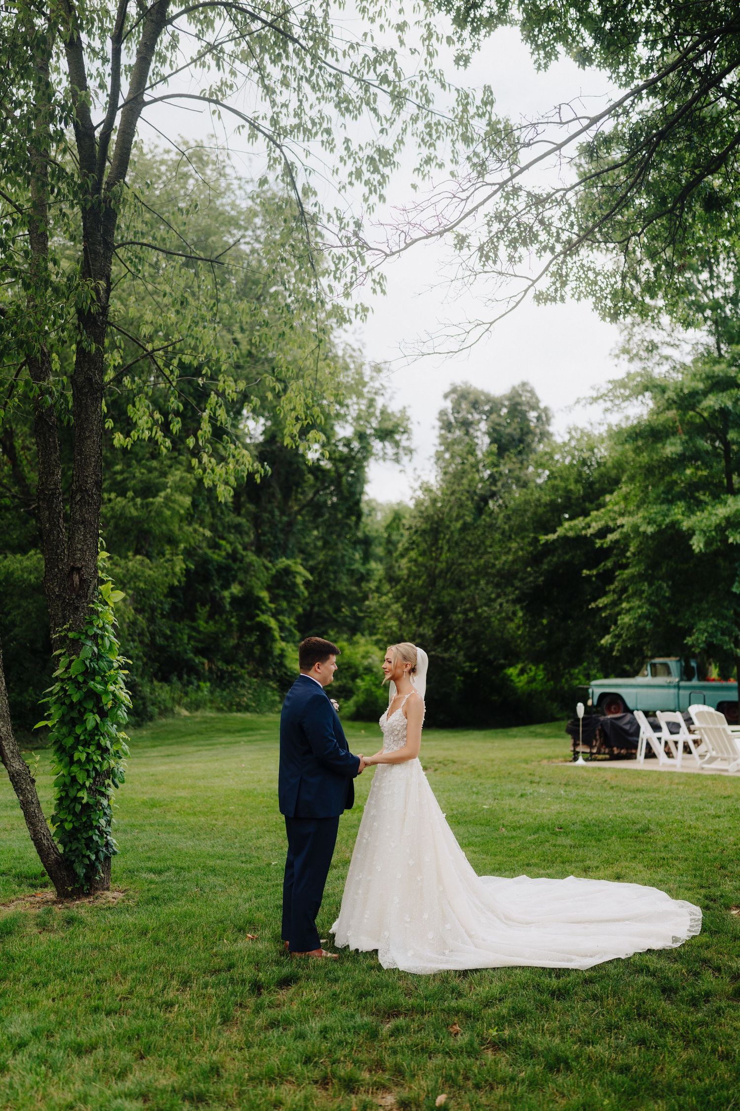 A wedding couple stands in a lush green garden setting with a flowing white dress train.