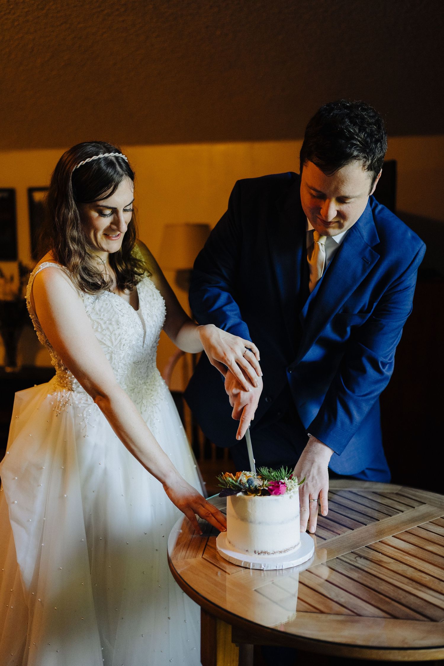 A couple in wedding attire cutting their small white wedding cake together on a wooden table.