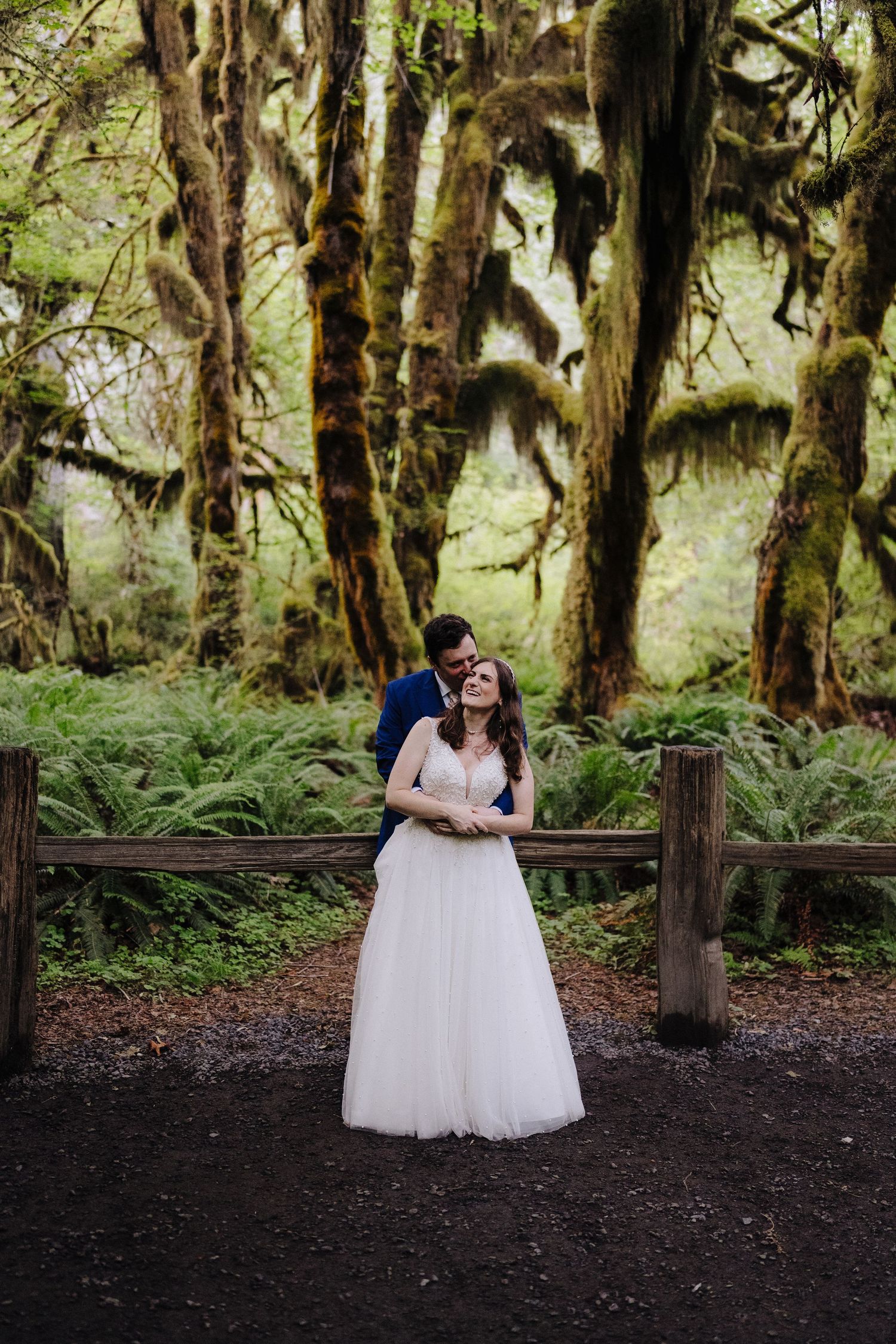 A couple embraces in a lush forest setting with moss-covered trees and ferns.
