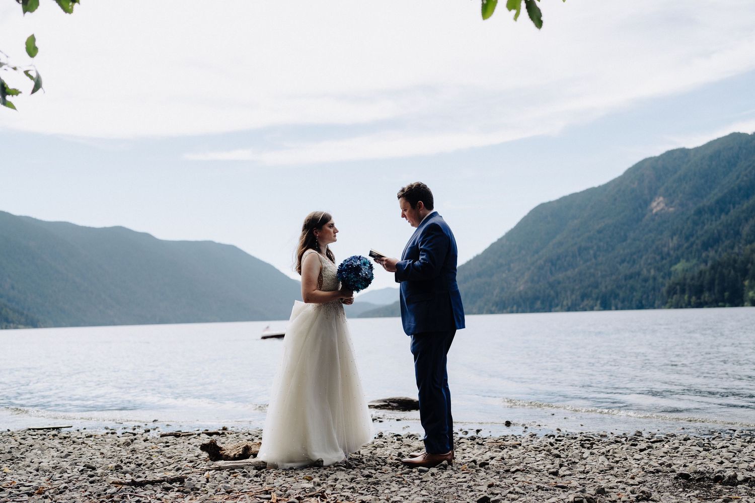 A romantic wedding scene unfolds on a pebble beach with mountains and water creating a stunning backdrop.